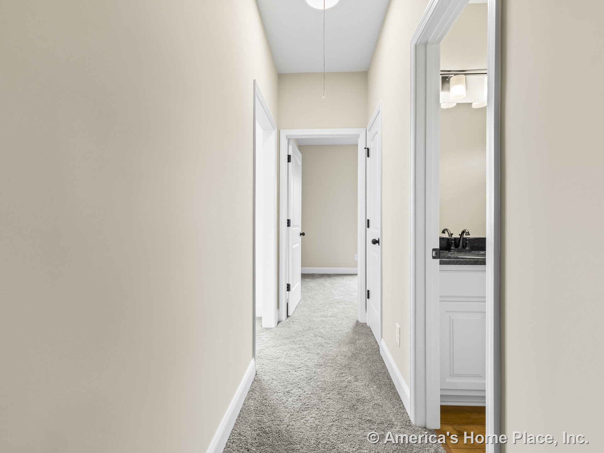 Carpeted hallway with white interior doors and trim, leading to a bathroom featuring a built-in vanity with granite countertop, neutral wall color, ceiling light fixture, and white