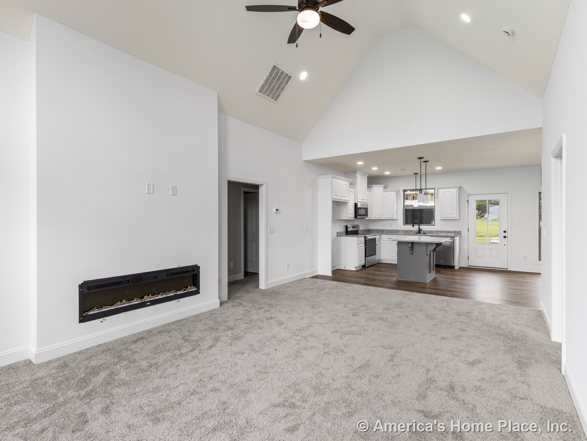 Vaulted ceiling with recessed lighting above open living area, built-in electric fireplace set into wall, carpeted living room adjacent to kitchen with white cabinetry, granite