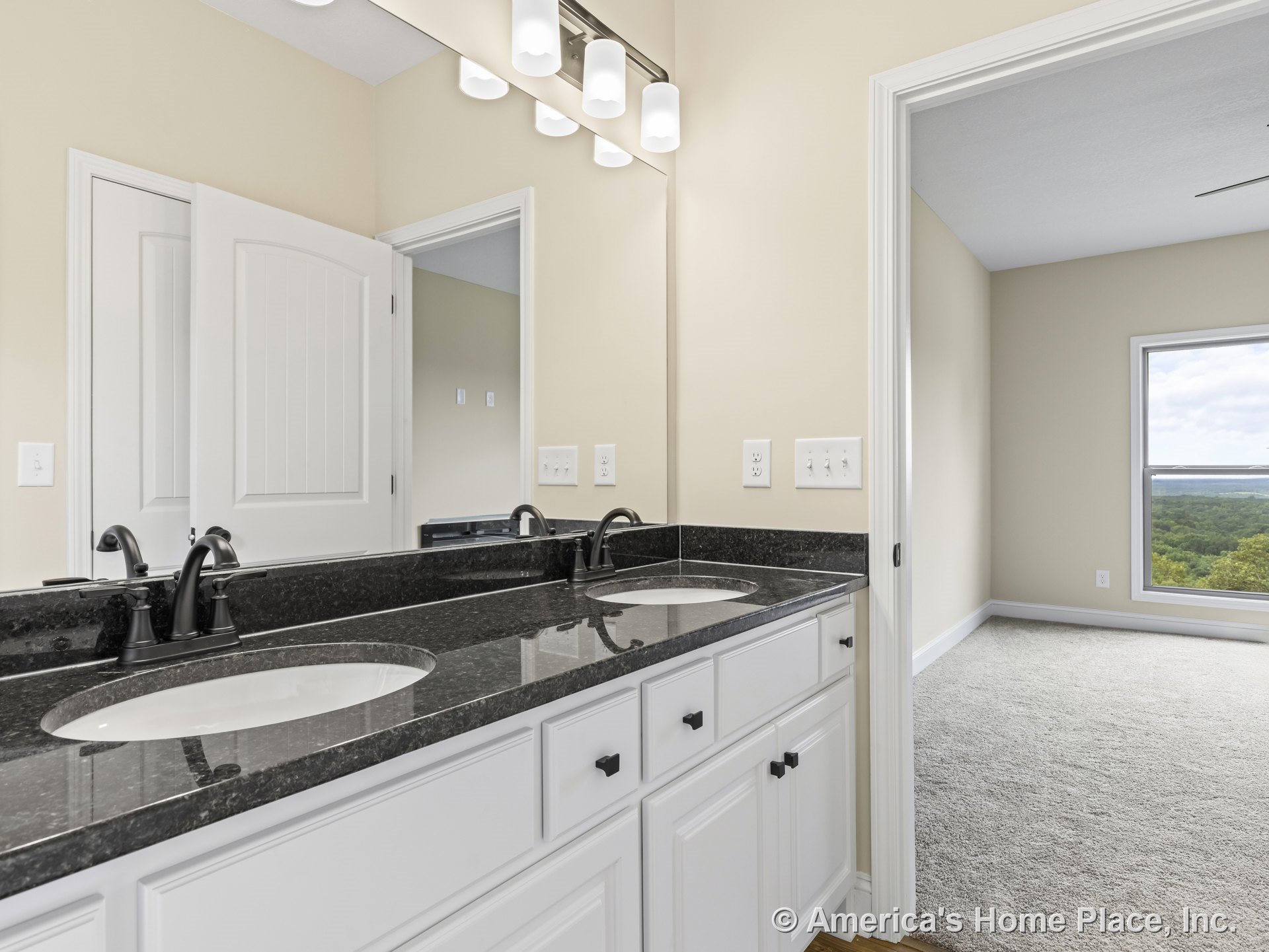 Double vanity with black granite countertop, white shaker cabinets, matte black faucets, wall-mounted light fixtures above large mirrors, panel interior door, view into adjacent