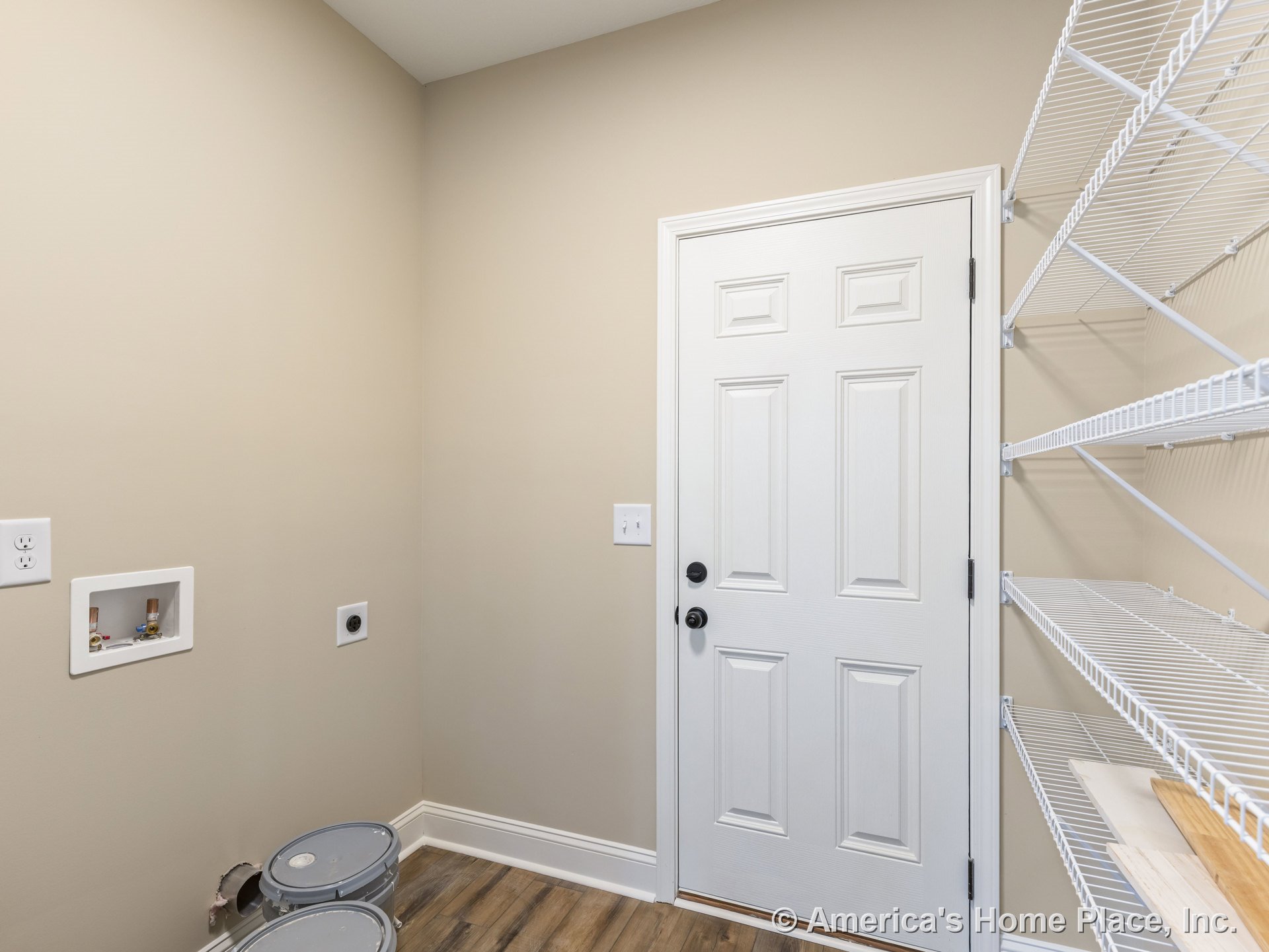 Laundry room with built-in white wire shelving, wall-mounted utility hookups, paneled exterior door, beige painted walls, wood-look flooring, and white baseboard trim.