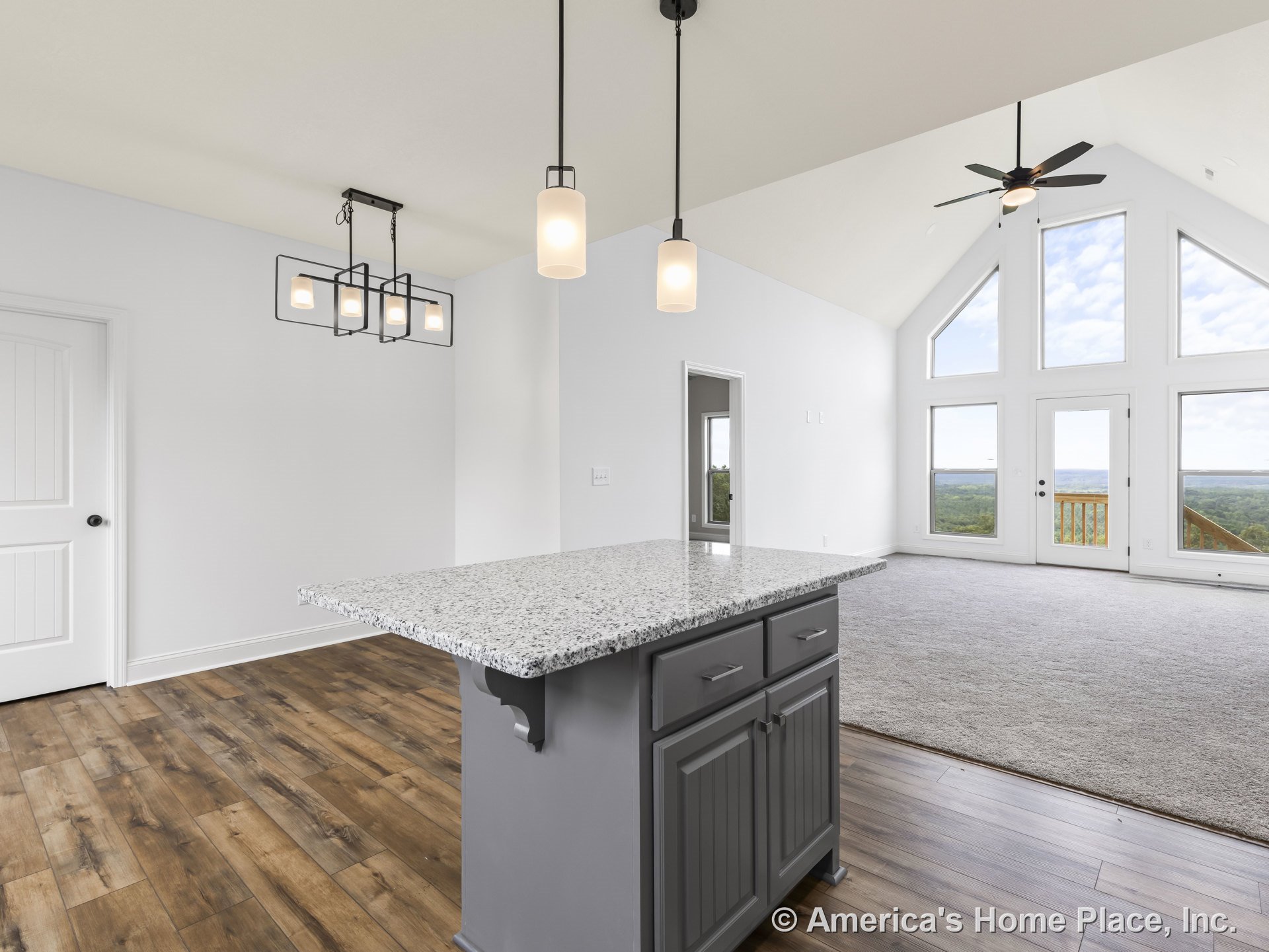 Open concept living area with vaulted ceiling and fan, large triangular window wall, granite kitchen island, modern pendant lights, wood-look flooring, and white interior trim.