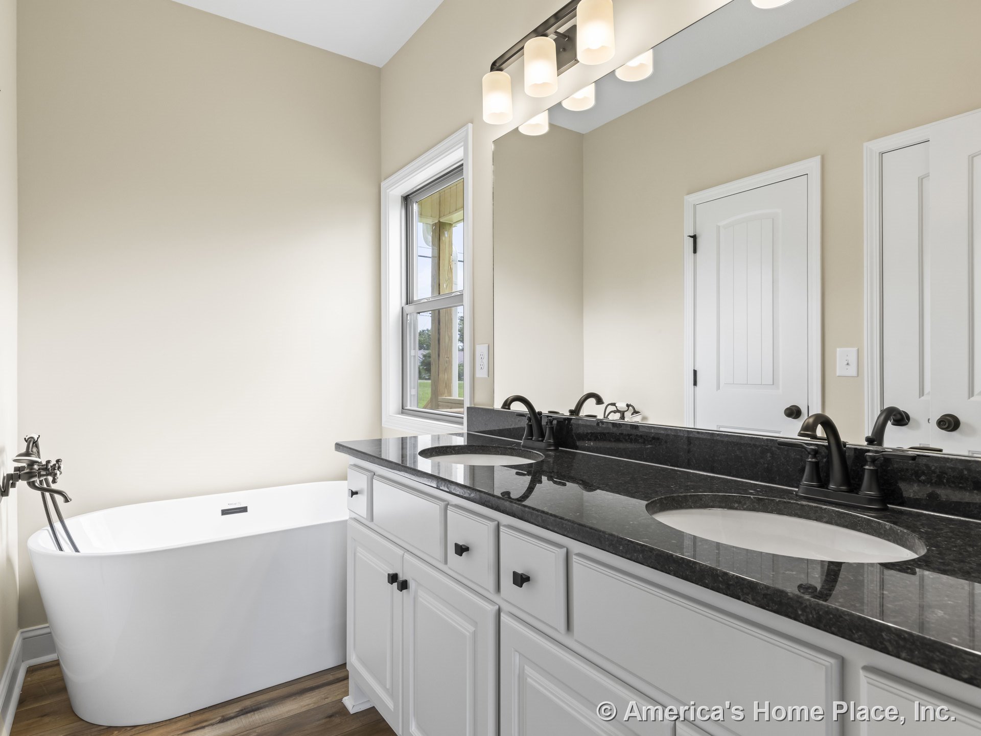 Freestanding soaking tub beside a double sink vanity with black granite countertop, matte black faucets, white cabinetry, wall-mounted light fixtures above a large mirror, wood