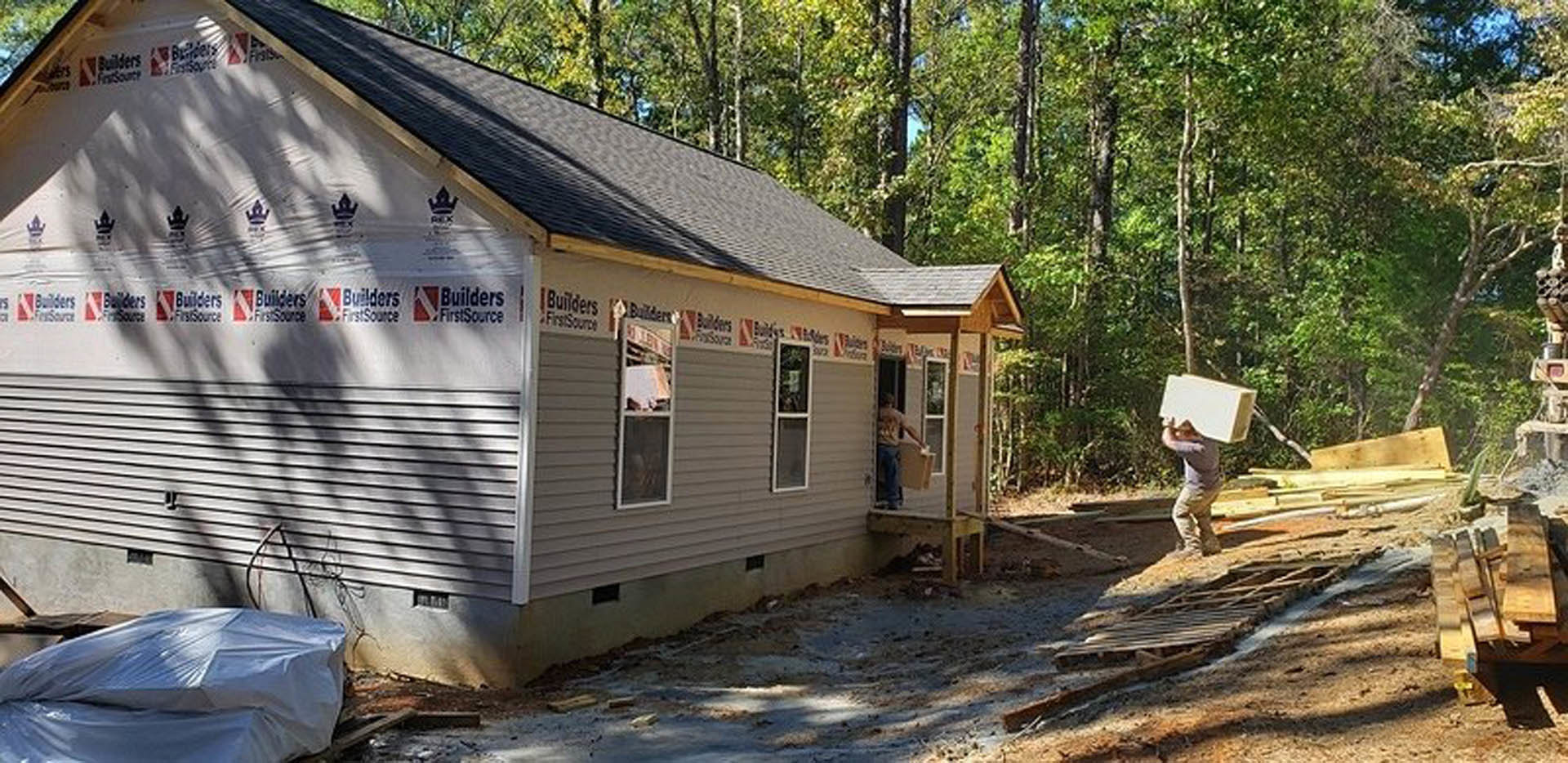 White siding custom home with gabled roof, wood plank porch, mature trees, and a man holding papers and a bag standing near the entrance