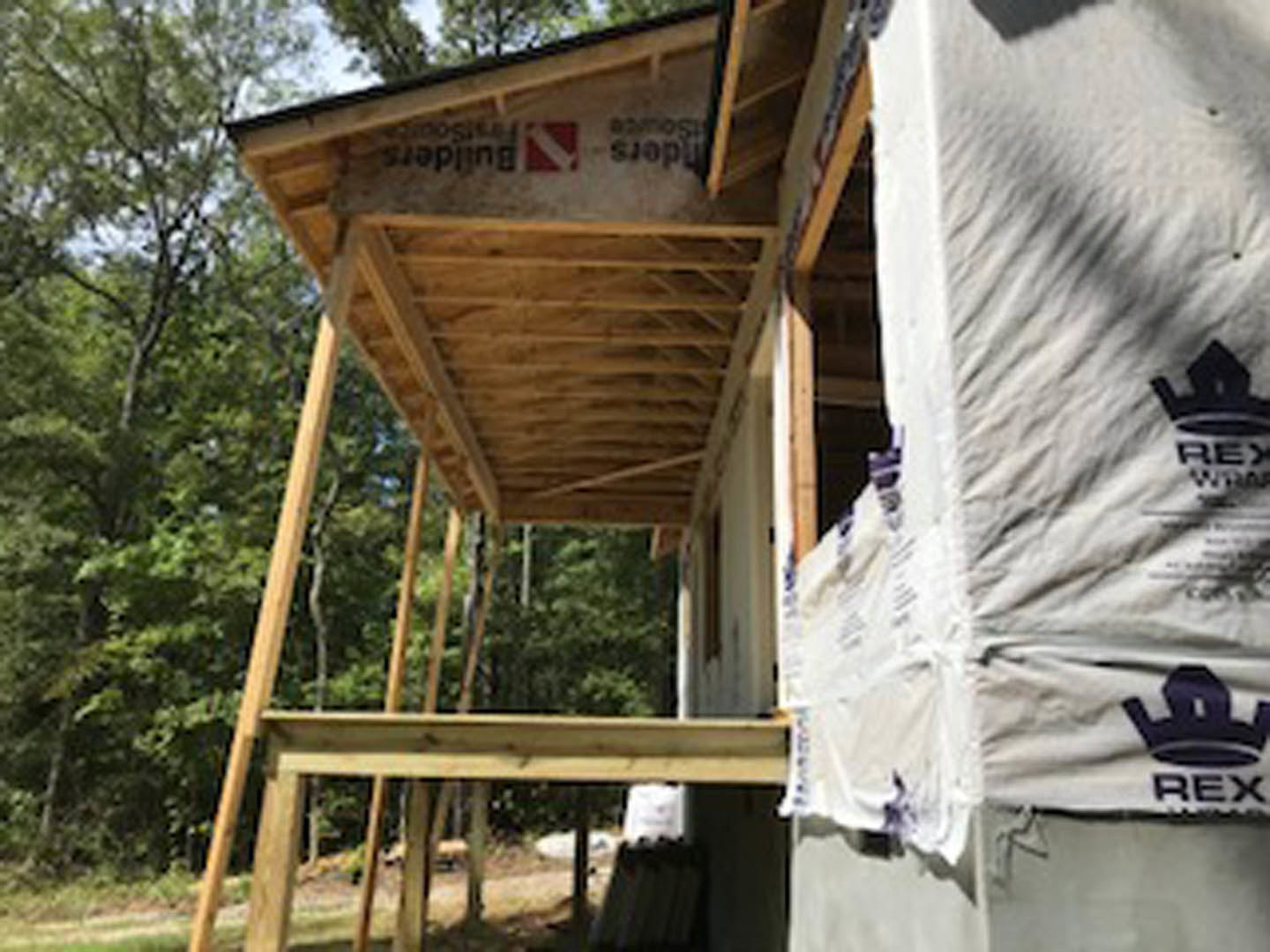 Partially built wooden house with exposed framing, covered porch, and roof; construction materials scattered in front; leafy trees in background.