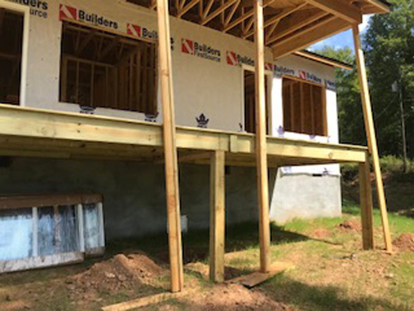 Partially built house with exposed wooden beams, unfinished roof, and several installed windows; grassy yard and trees in background.