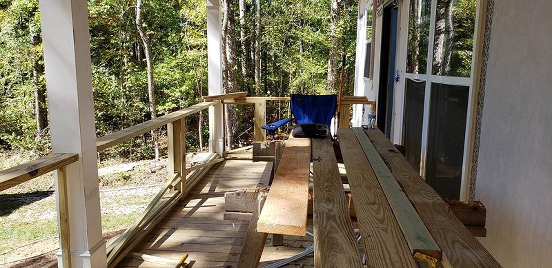 Wood deck with outdoor dining table and blue cushioned chairs, surrounded by trees and greenery, wooden handrail visible along edge