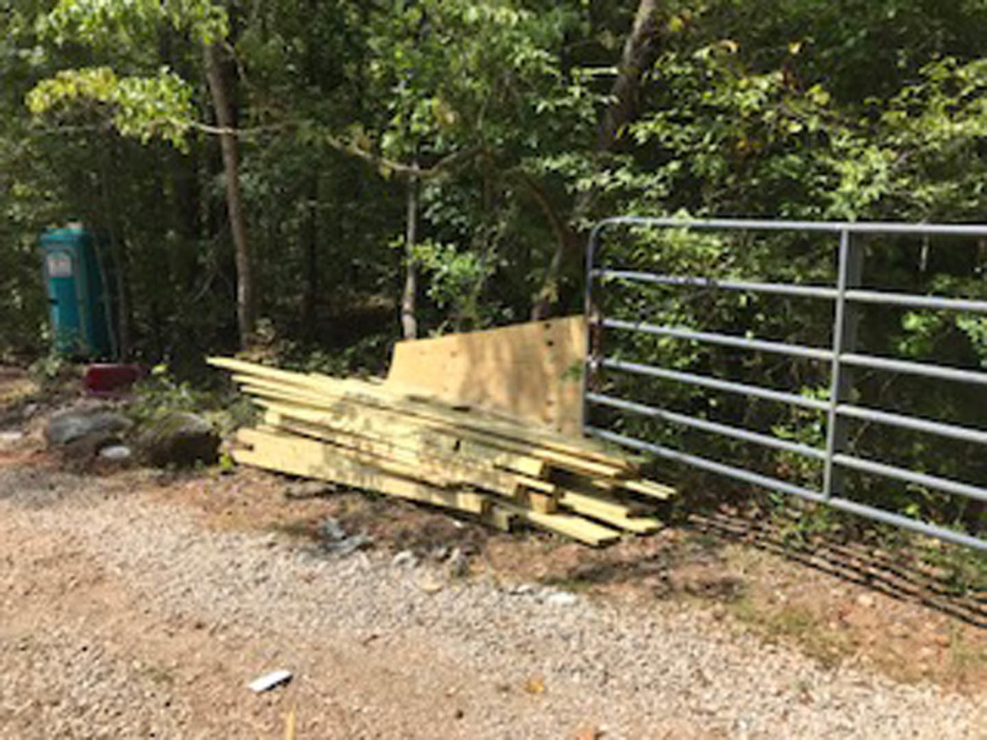 Stacked lumber beside a metal gate secured with a chain and padlock, set on grassy ground near a stone and surrounded by outdoor plants and fencing.