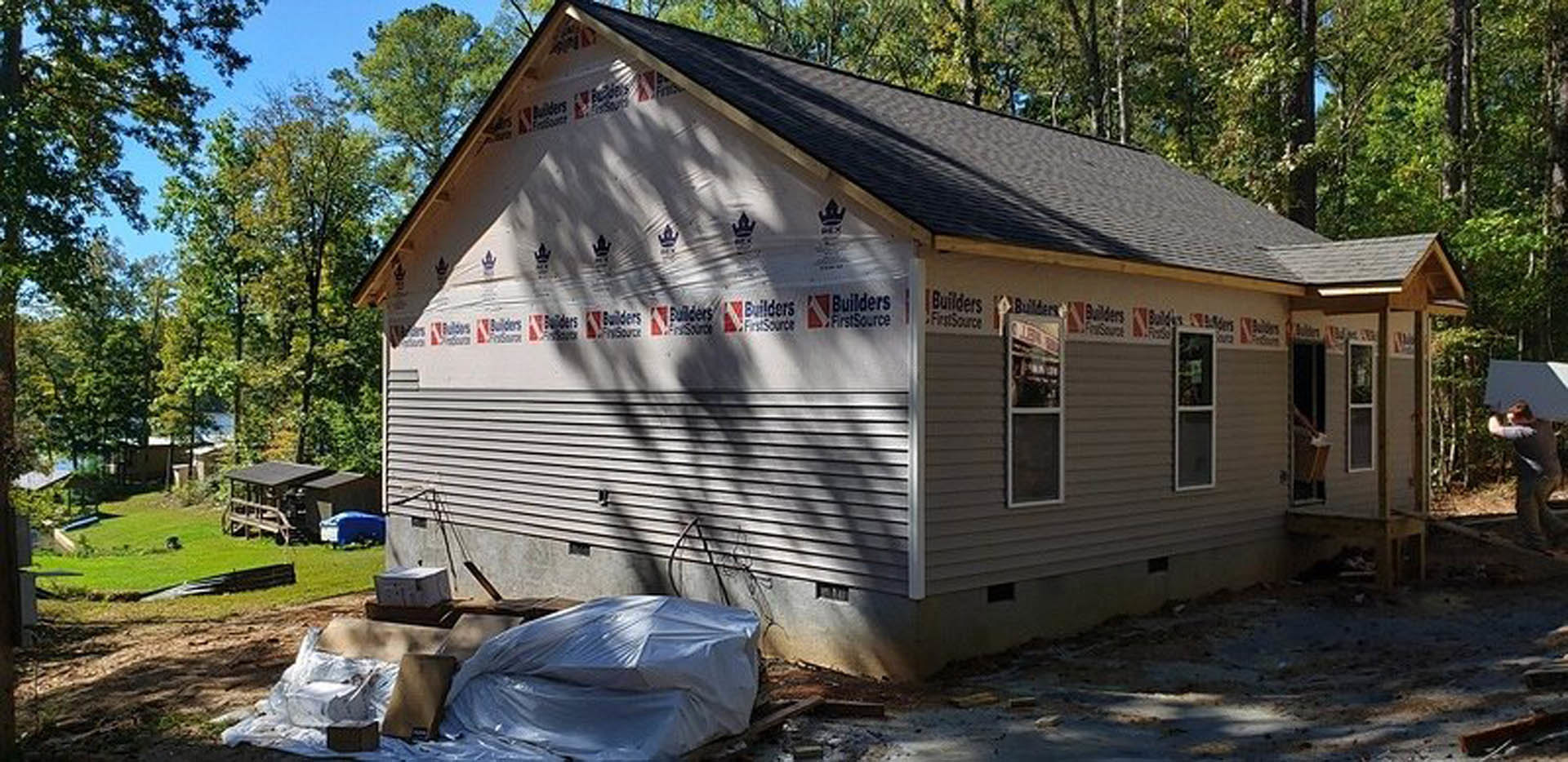 Wood-framed house under construction with exposed beams, unfinished siding, and leafy trees in the background