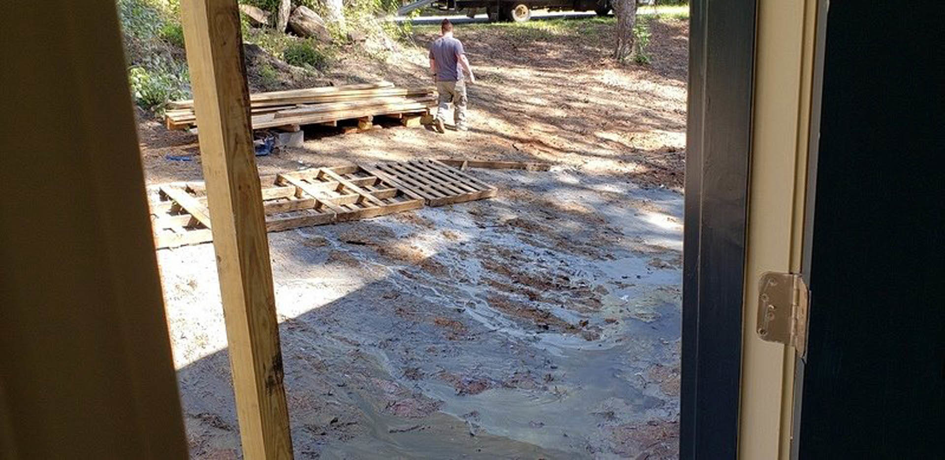 Man standing on muddy ground beside a stack of wooden boards outside a custom home, with trees and windows visible in the background
