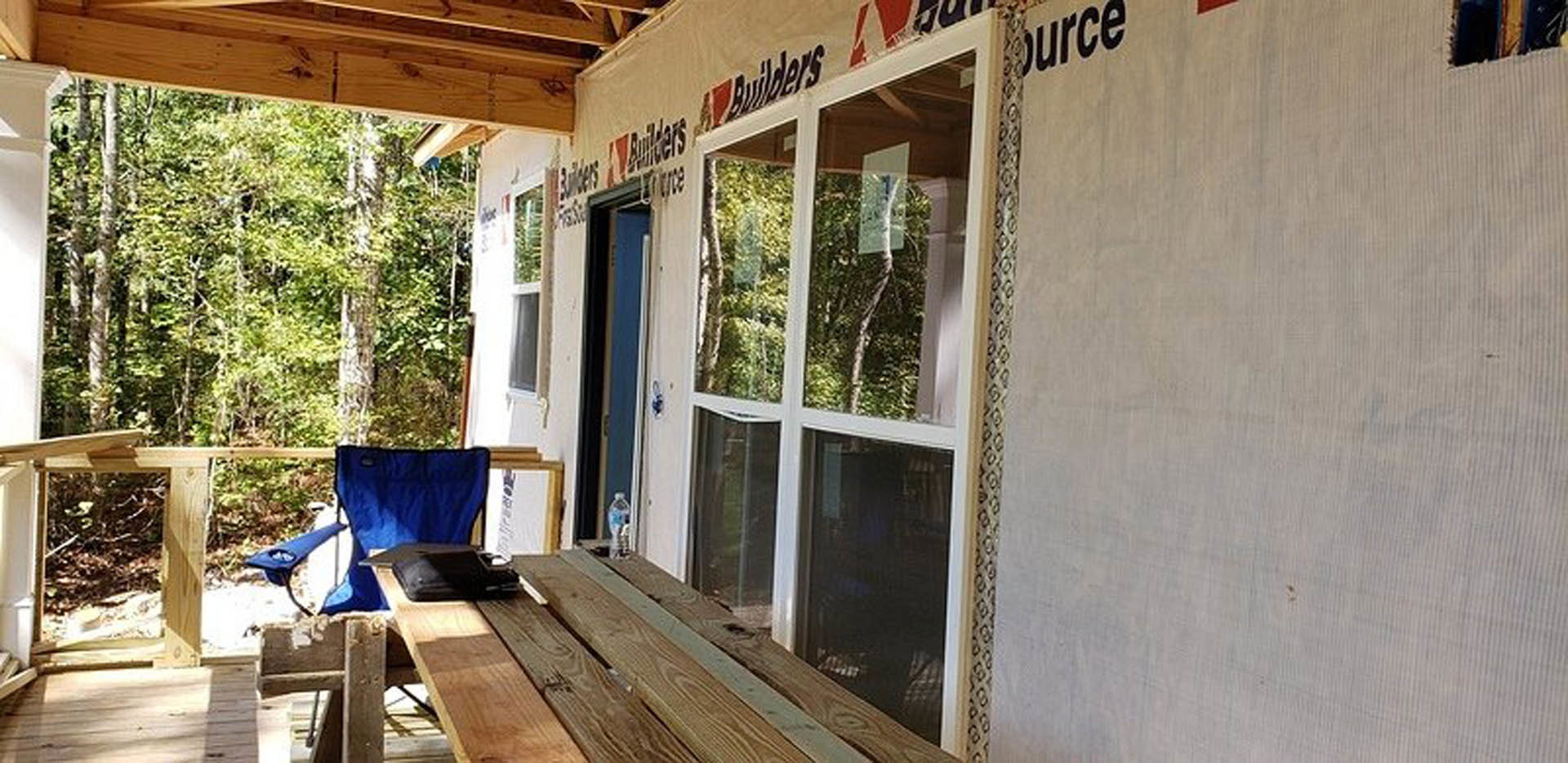 Wood table and blue chair on shaded outdoor porch with trees and plants, building exterior features wood plank siding and large windows.