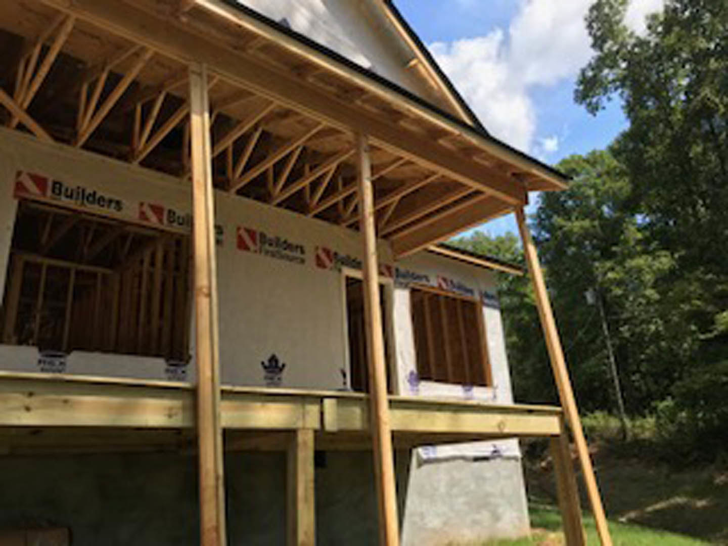Wood-framed custom home under construction with exposed beams, partially finished roof, surrounding trees, and cloudy sky in the background