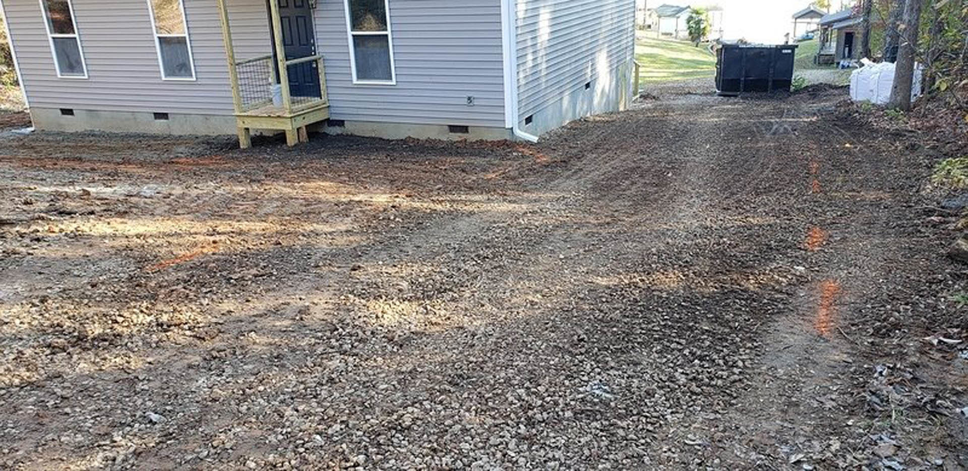 Dirt driveway leading to a wooden house with a front door, windows, surrounding trees, and a dog standing near the entrance