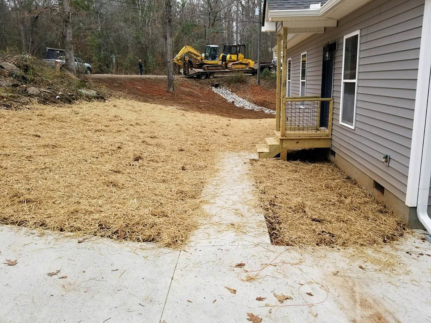 Modern home with large windows, brown siding, and a landscaped yard featuring a white stone border; yellow bulldozer parked on a flatbed trailer near a tree and a car.