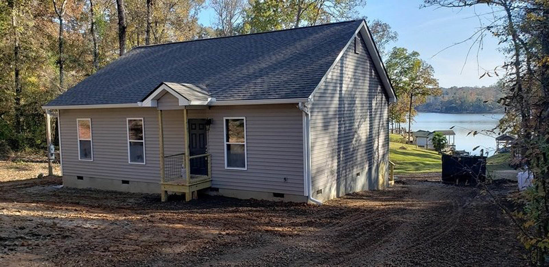 Two-story home with white-framed windows, covered front porch, gray shingle roof, and dirt yard bordered by mature trees