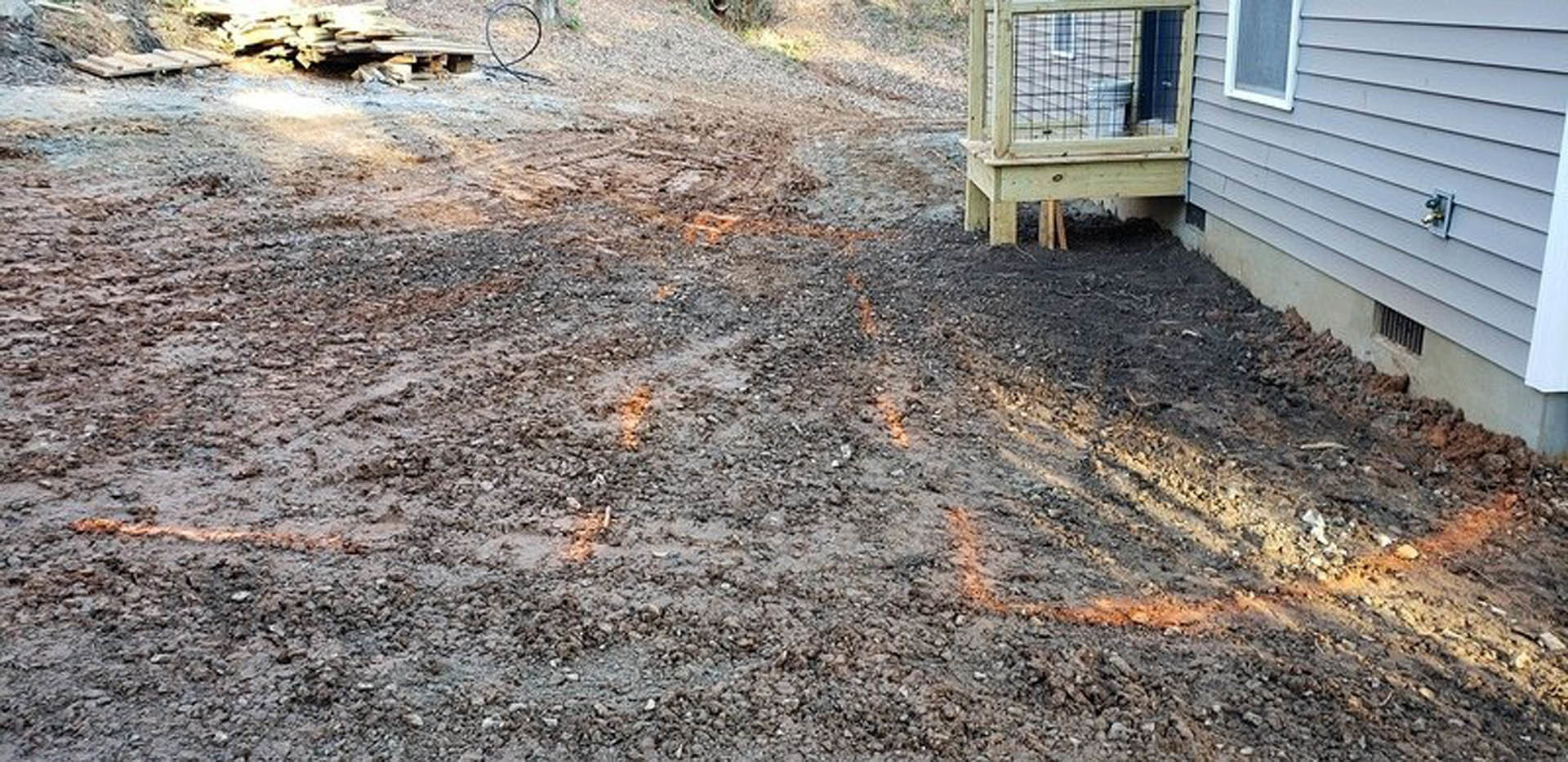 Dirt yard bordered by wooden fence, single-story house with wood siding, scattered grass patches, and mature trees in background