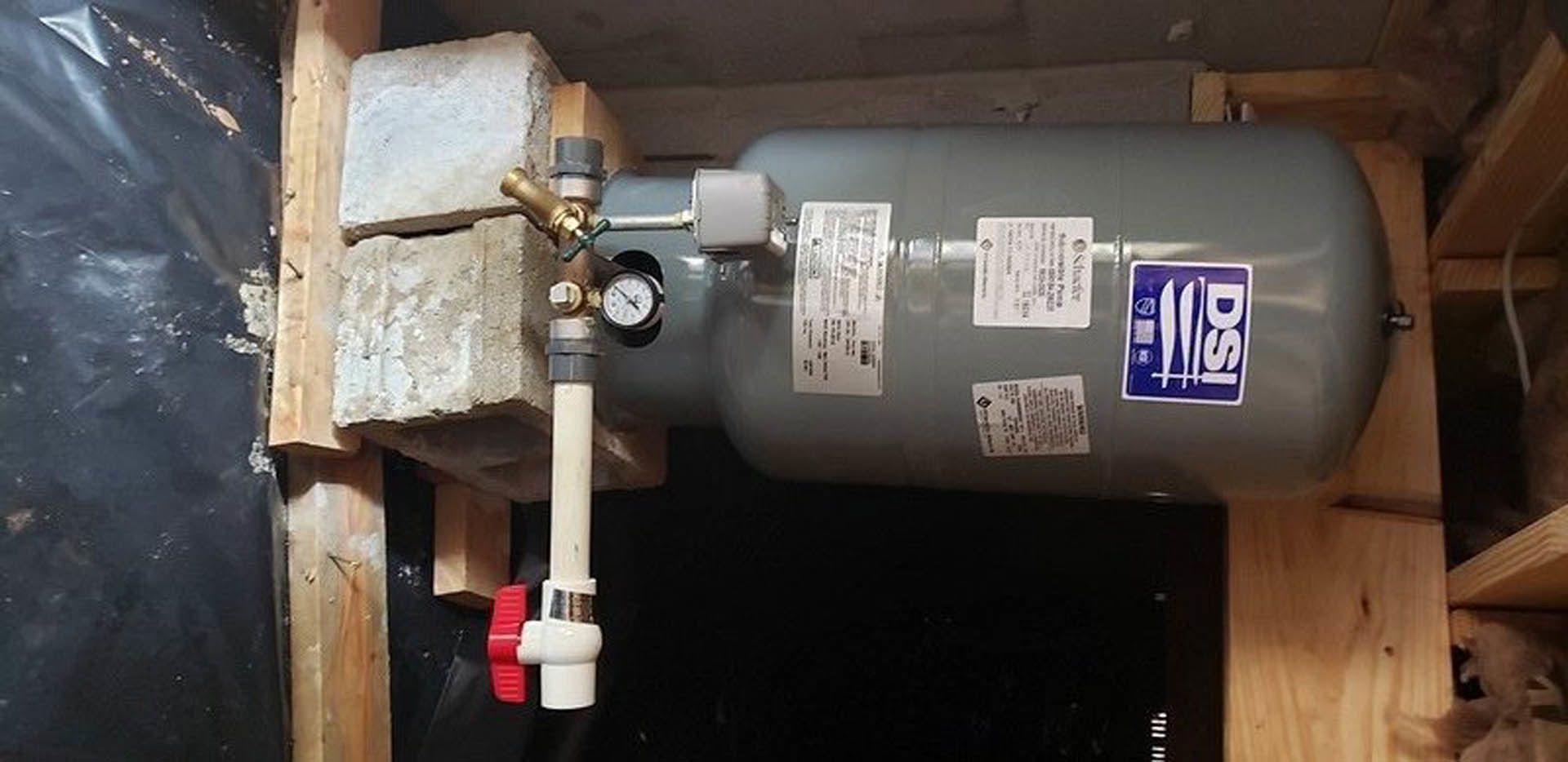 Grey metal gas cylinder with attached valve and white pipe, positioned indoors against a wall with visible labels.