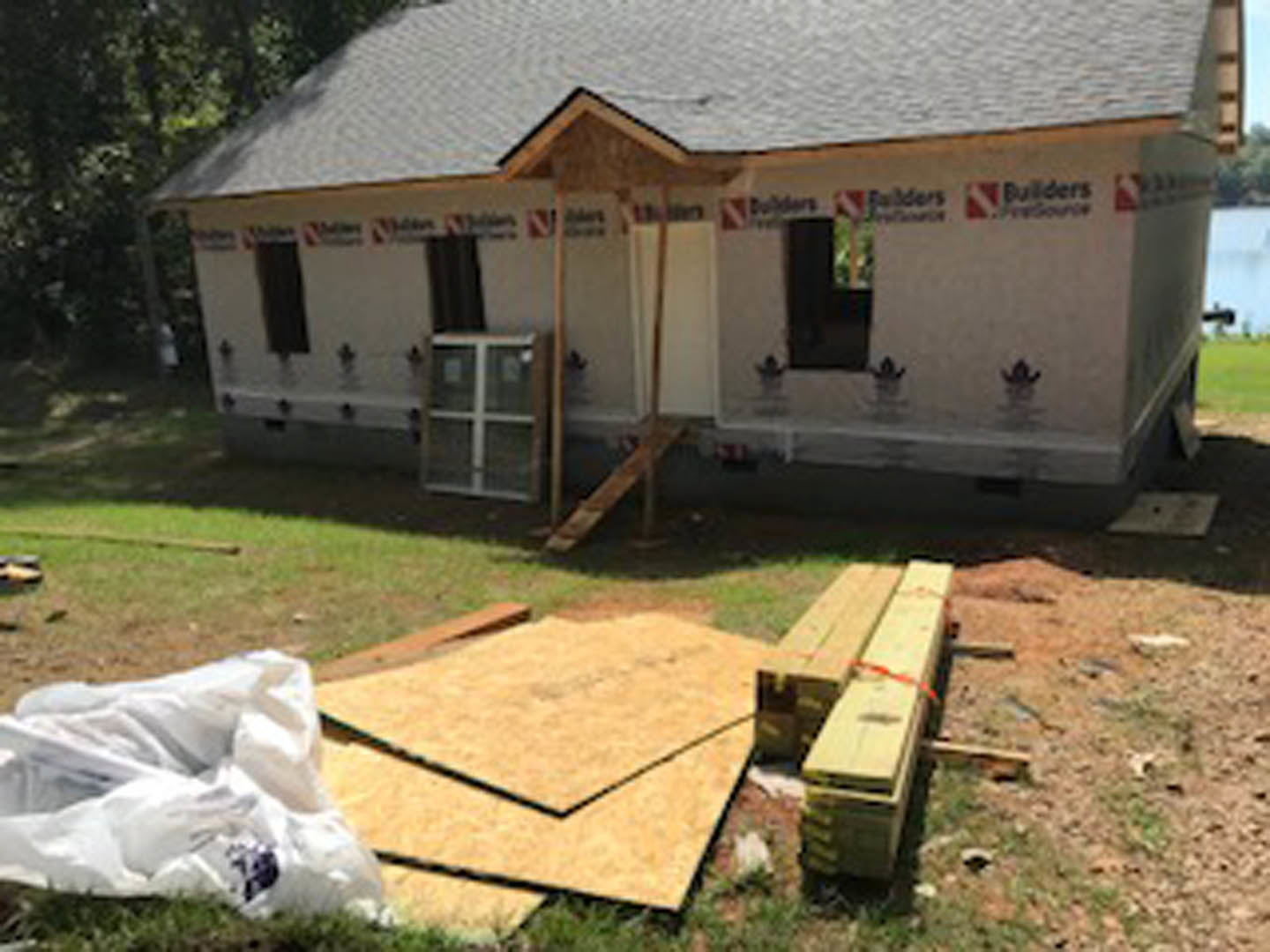 Wood-framed house under construction with exposed beams, stacked lumber on grassy yard, ladder leaning against unfinished exterior, trees and shed in background