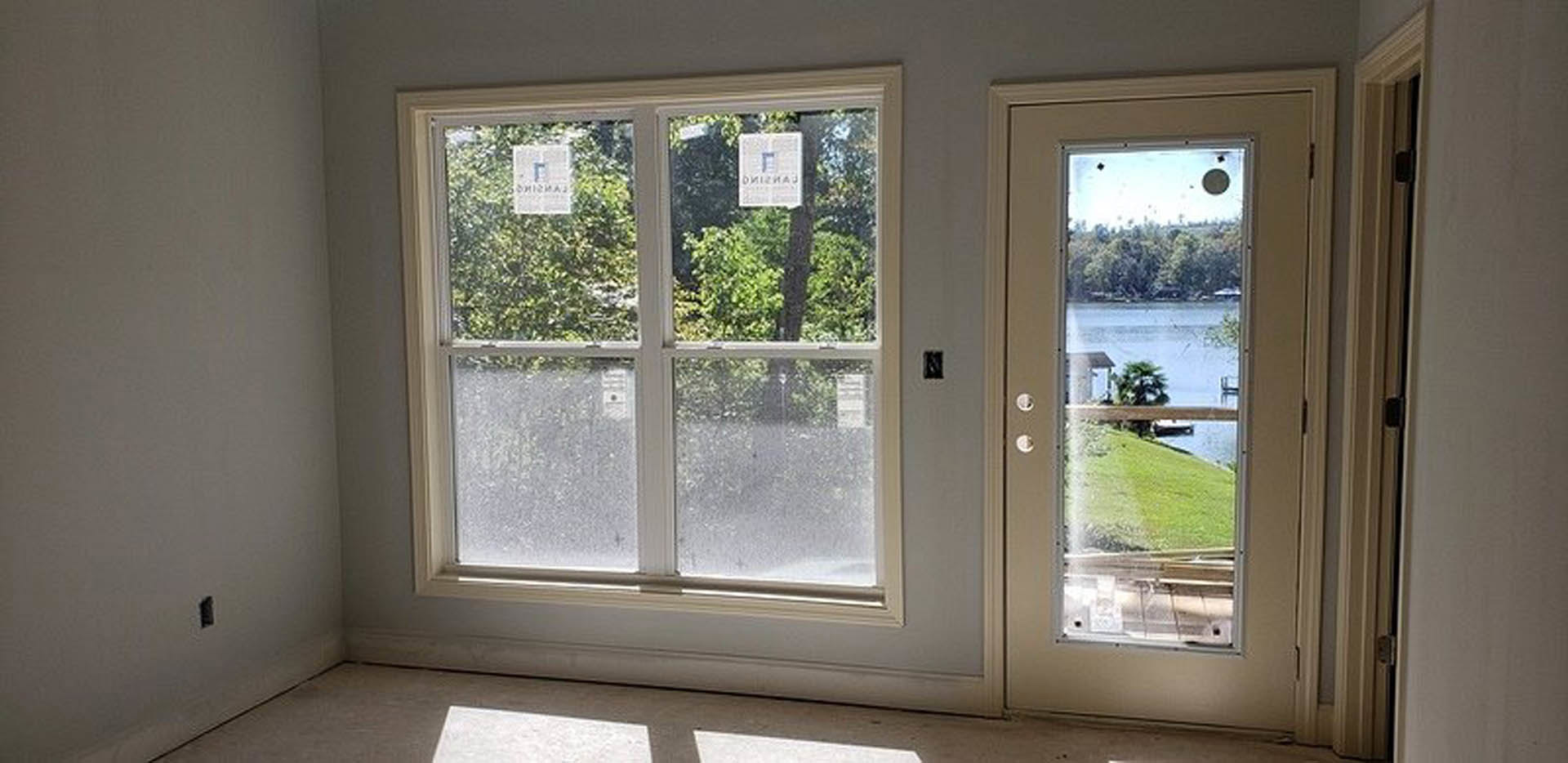 Room with large window and glass door overlooking lake, tree visible outside, window features signage, neutral walls and flooring