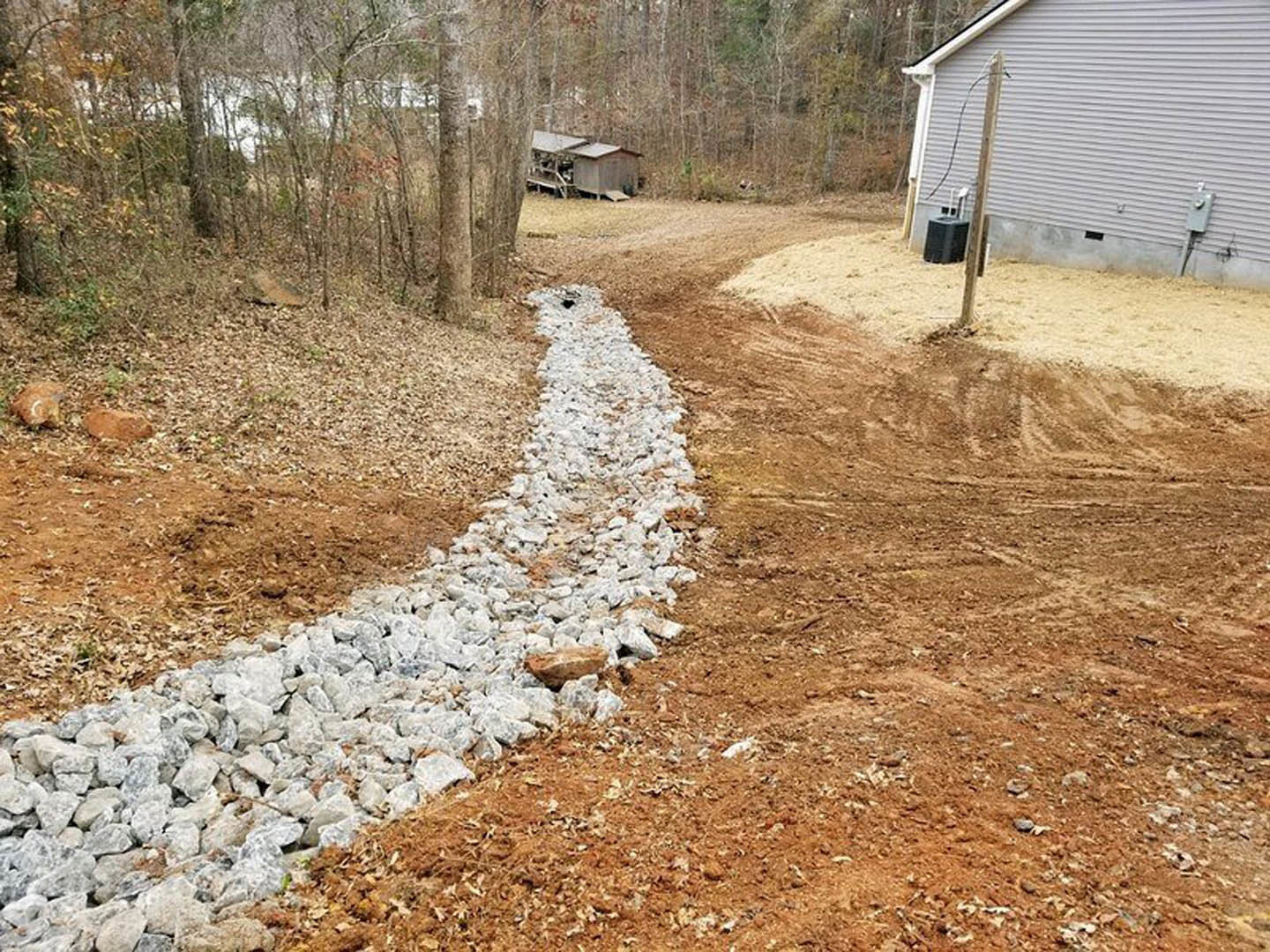 Gravel path bordered by rocks and dirt leading through a yard with trees, grey siding wall featuring electrical boxes, wooden shed nestled in the woods, and black and grey