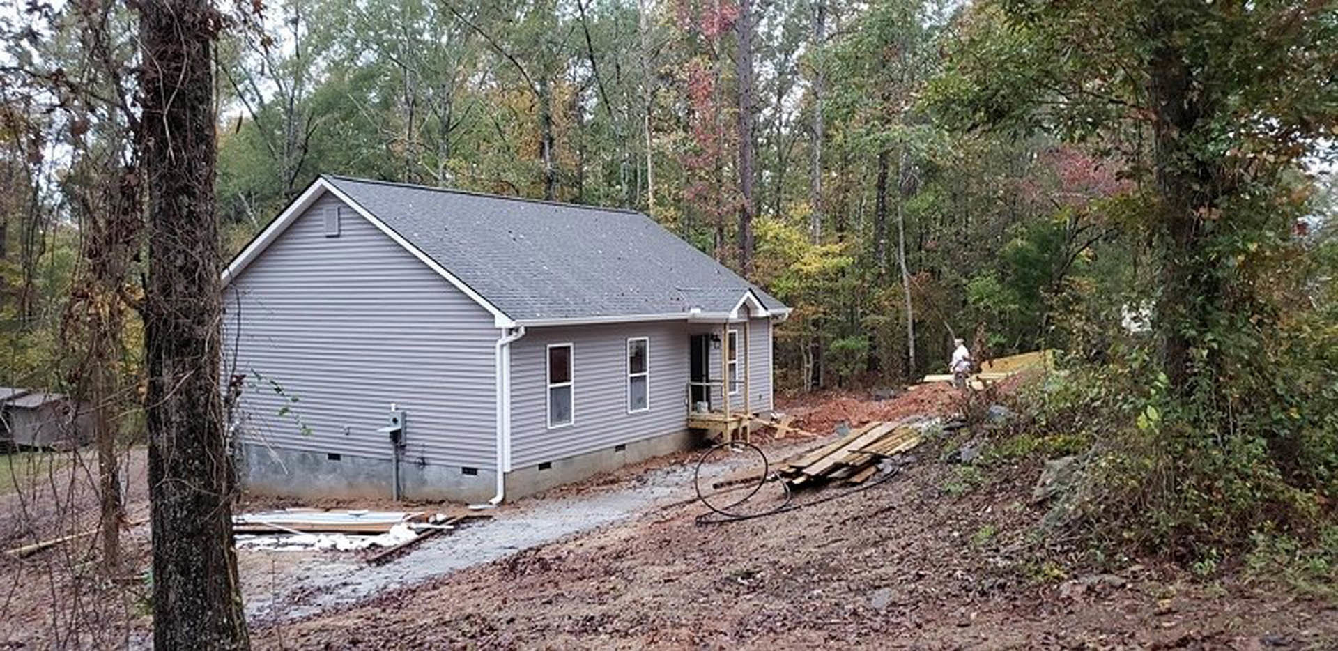 Wood-framed house under construction surrounded by tall trees, exposed siding and unfinished roof, dirt ground with scattered building materials