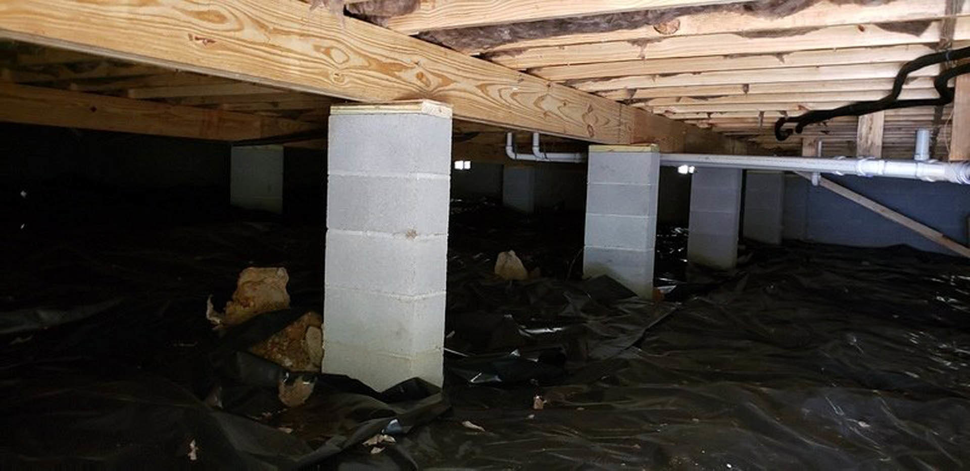 Unfinished basement with exposed wooden ceiling beams, concrete floor scattered with white blocks, black tarp along one wall, and visible insulation.
