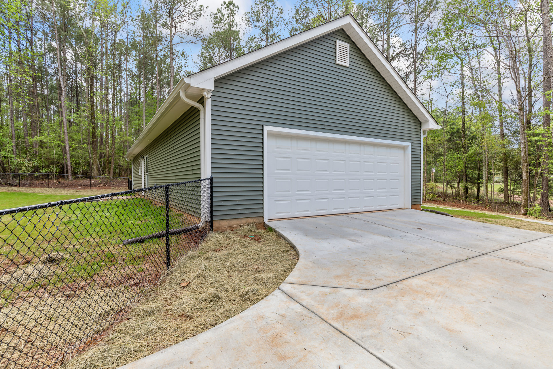 White garage door with gray siding, concrete driveway, wooden fence, green grass, and trees under a clear sky