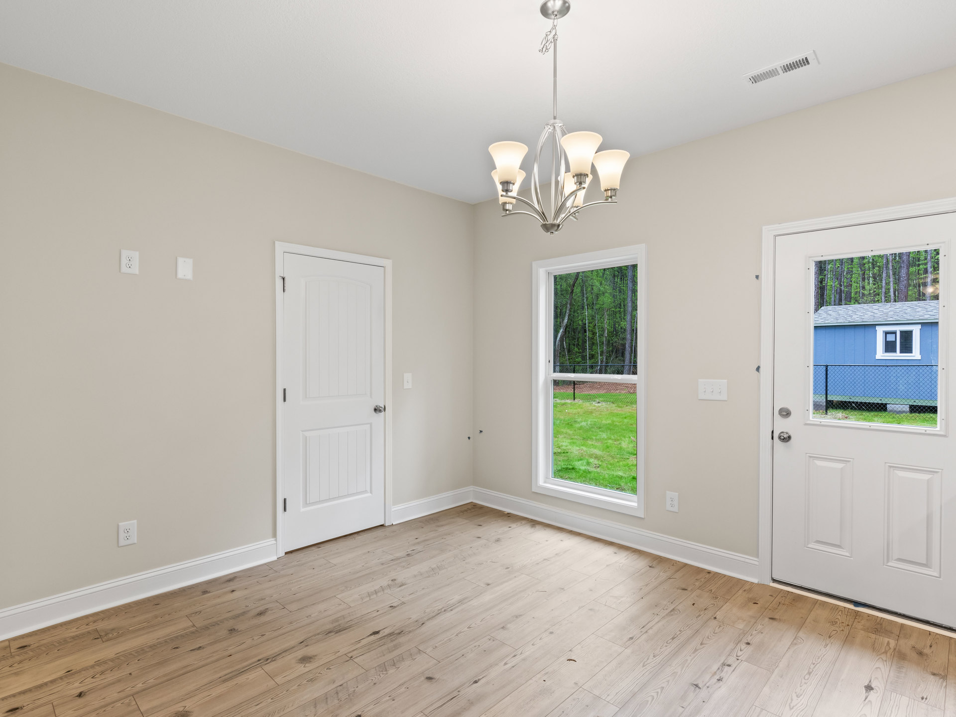 Chandelier with four white shades hanging in a room featuring wood flooring, white trim, plaster walls, and two white doors—one with a silver knob, the other with a window and view