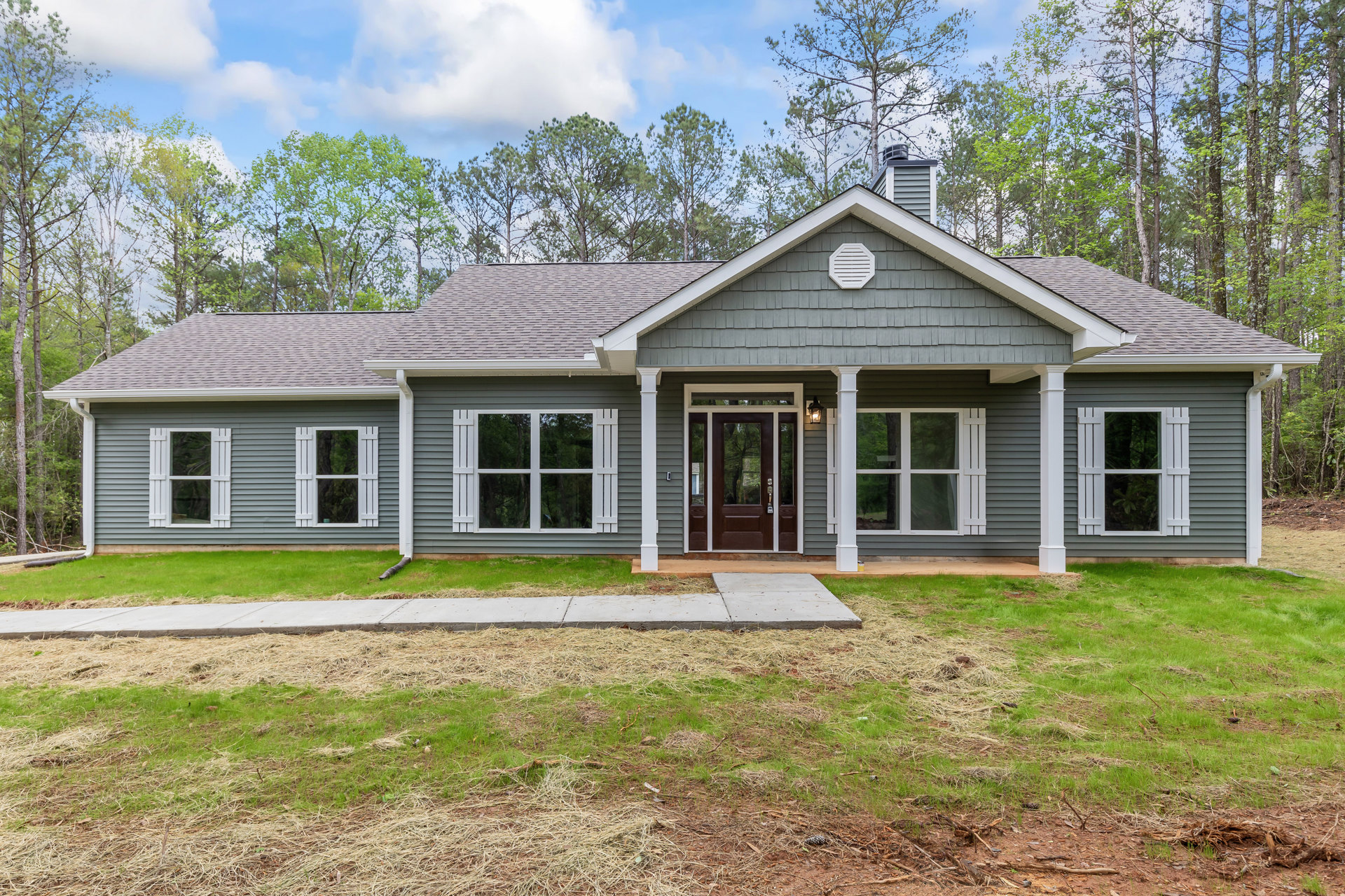 Gray custom home with white trim, brown glass-paneled front door, covered porch, white vent, windows with white shutters, manicured lawn, mature trees, and a smaller white house