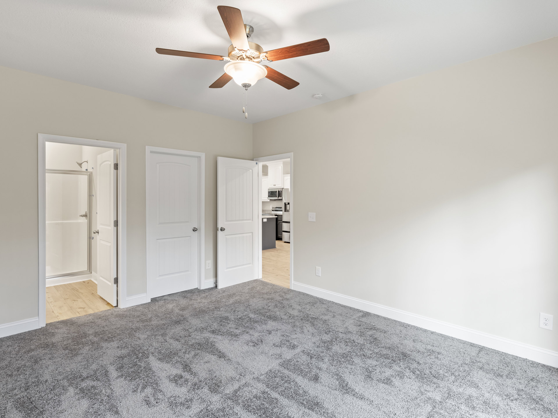 Bedroom with grey carpet, white walls, ceiling fan with light fixture, white door with silver knob, open glass shower door in adjoining bathroom