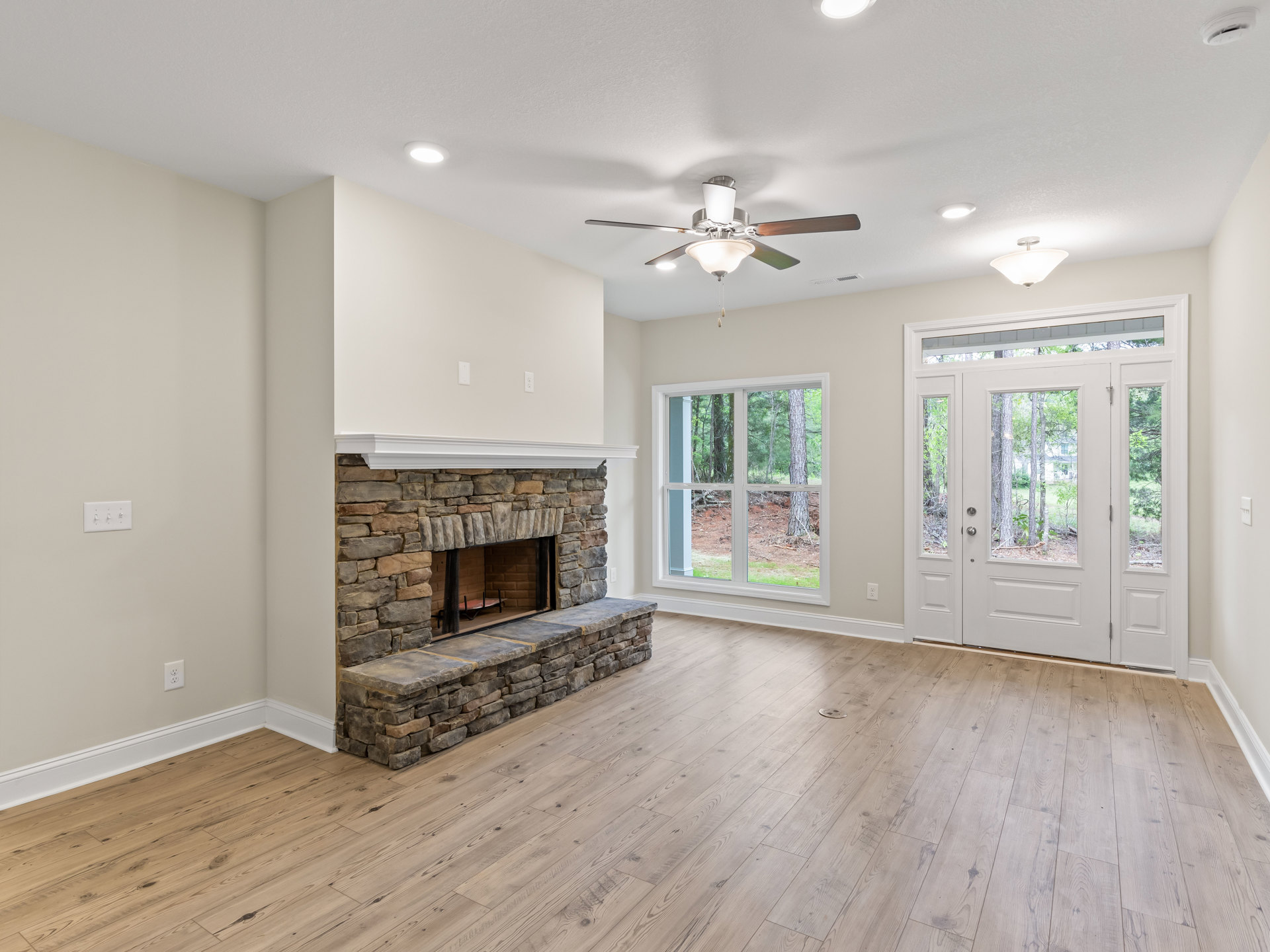 Living room with wood flooring, white fireplace mantel, ceiling fan with light, glass-paneled white door, and window overlooking trees
