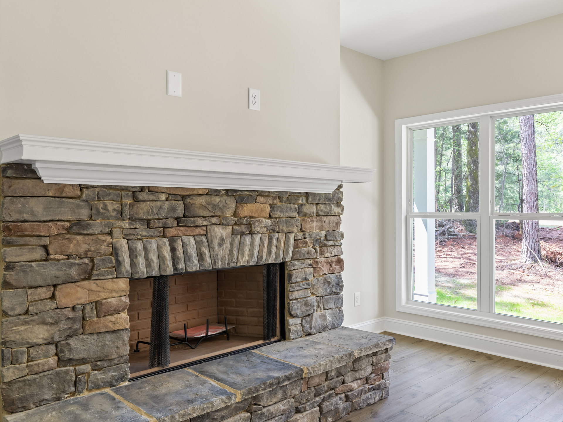 Stone fireplace with hearth and fire screen, flanked by a window showing trees outside, metal rack with plastic bag on stone steps, neutral walls and indoor finishes.