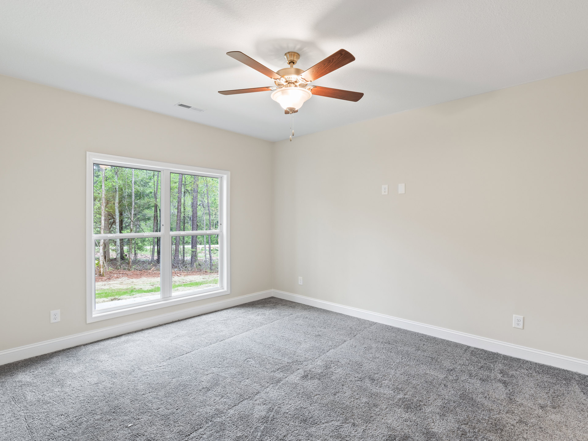 Neutral-toned bedroom with grey carpet, white walls, ceiling fan with light fixture, large window overlooking trees, simple crown molding