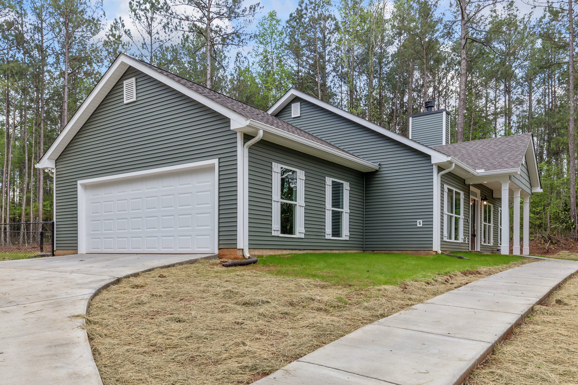 Two-story house with gray siding, white garage door, concrete driveway, and green grass lawn; white-framed windows with blinds; wooden fence bordering wooded area in background.