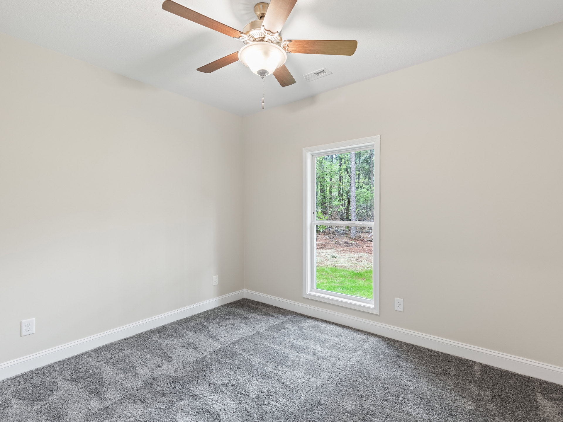Ceiling fan with light fixture mounted on white plaster ceiling, grey carpeted floor, large window revealing leafy trees outside
