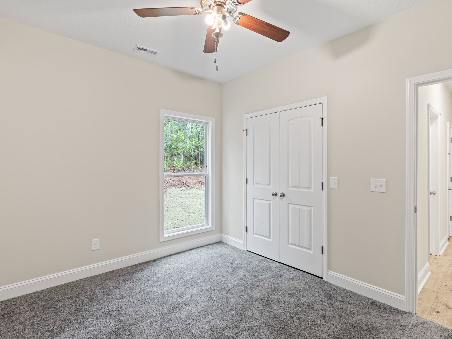 Bedroom with beige carpet, white closet doors with silver knobs, ceiling fan with lights, and window overlooking green trees.