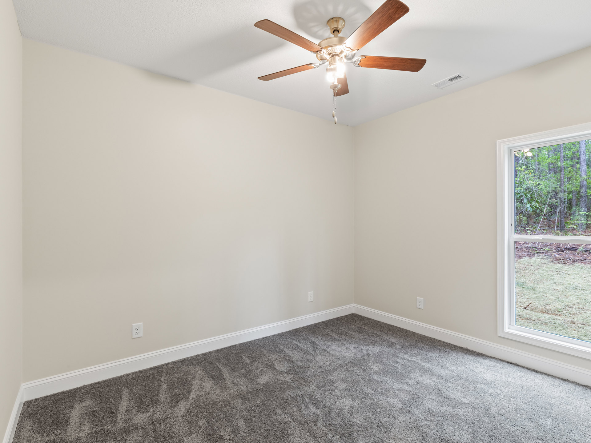 Ceiling fan with light fixture above grey carpeted floor, large window showing forest and grassy landscape, white plaster walls and ceiling