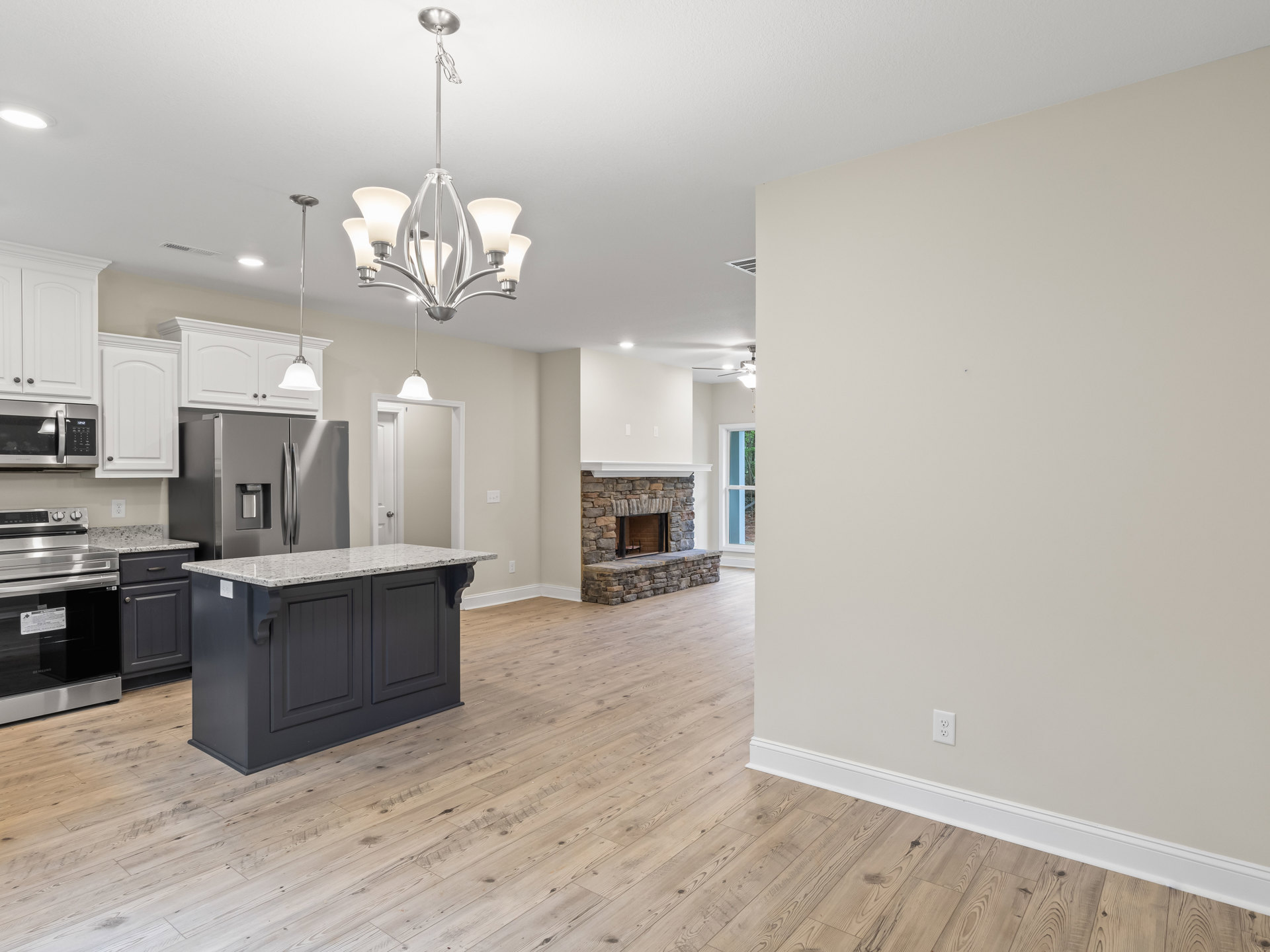 Open-concept kitchen and living room featuring a marble-topped island, stainless steel oven, white-shaded chandelier, built-in microwave, light wood cabinetry, stone fireplace, and