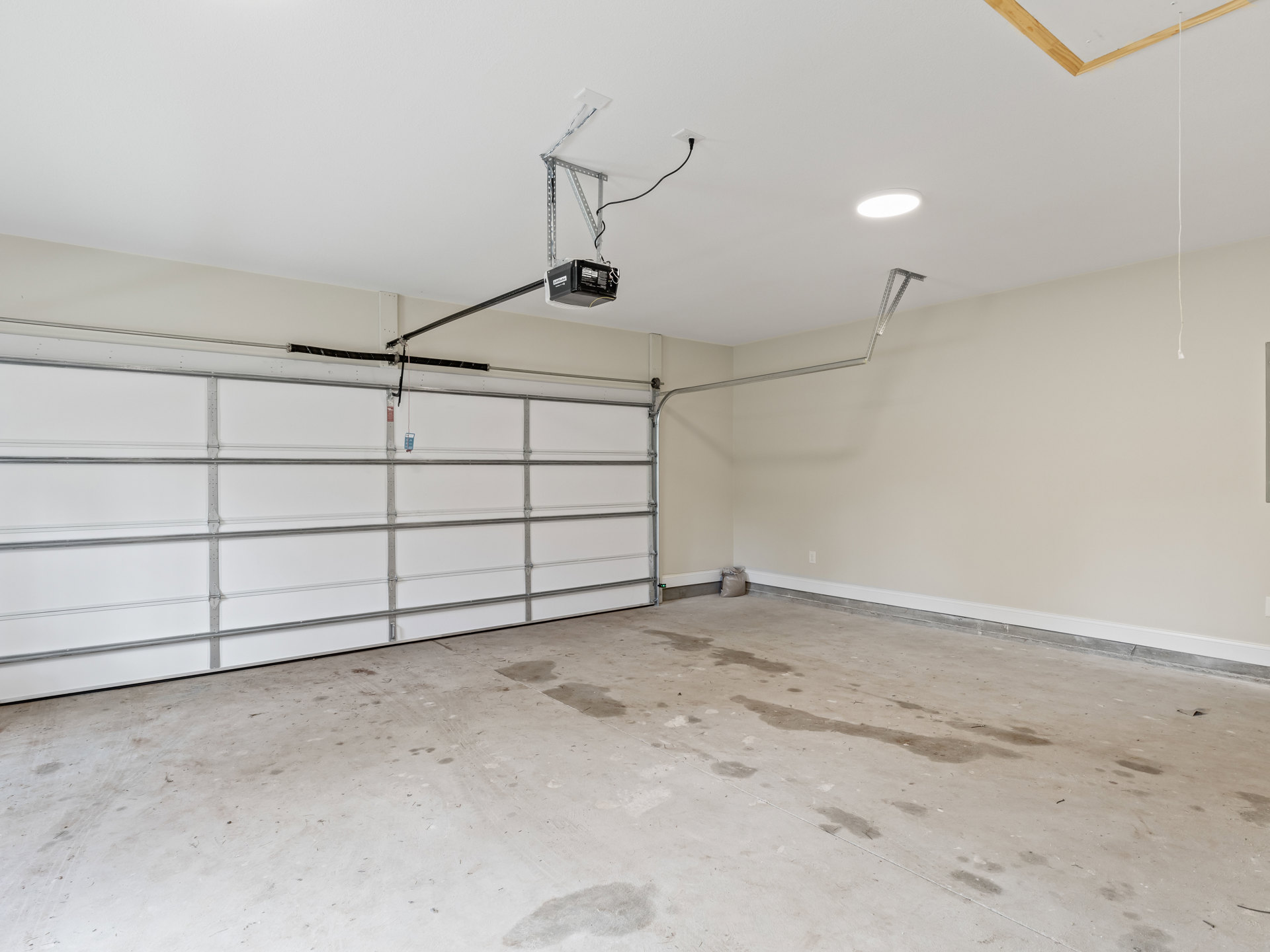 Garage interior with plaster walls, concrete floor, ceiling-mounted light fixture, metal-framed garage door, and a projector suspended from the ceiling; bag of water on the floor.