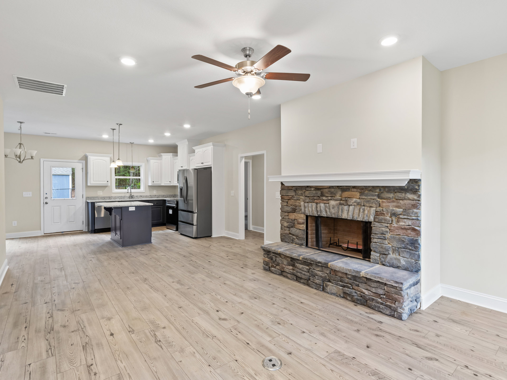 Open concept living room with stone fireplace, ceiling fan with light, adjacent kitchen featuring stainless steel refrigerator, wood flooring, and white door with window