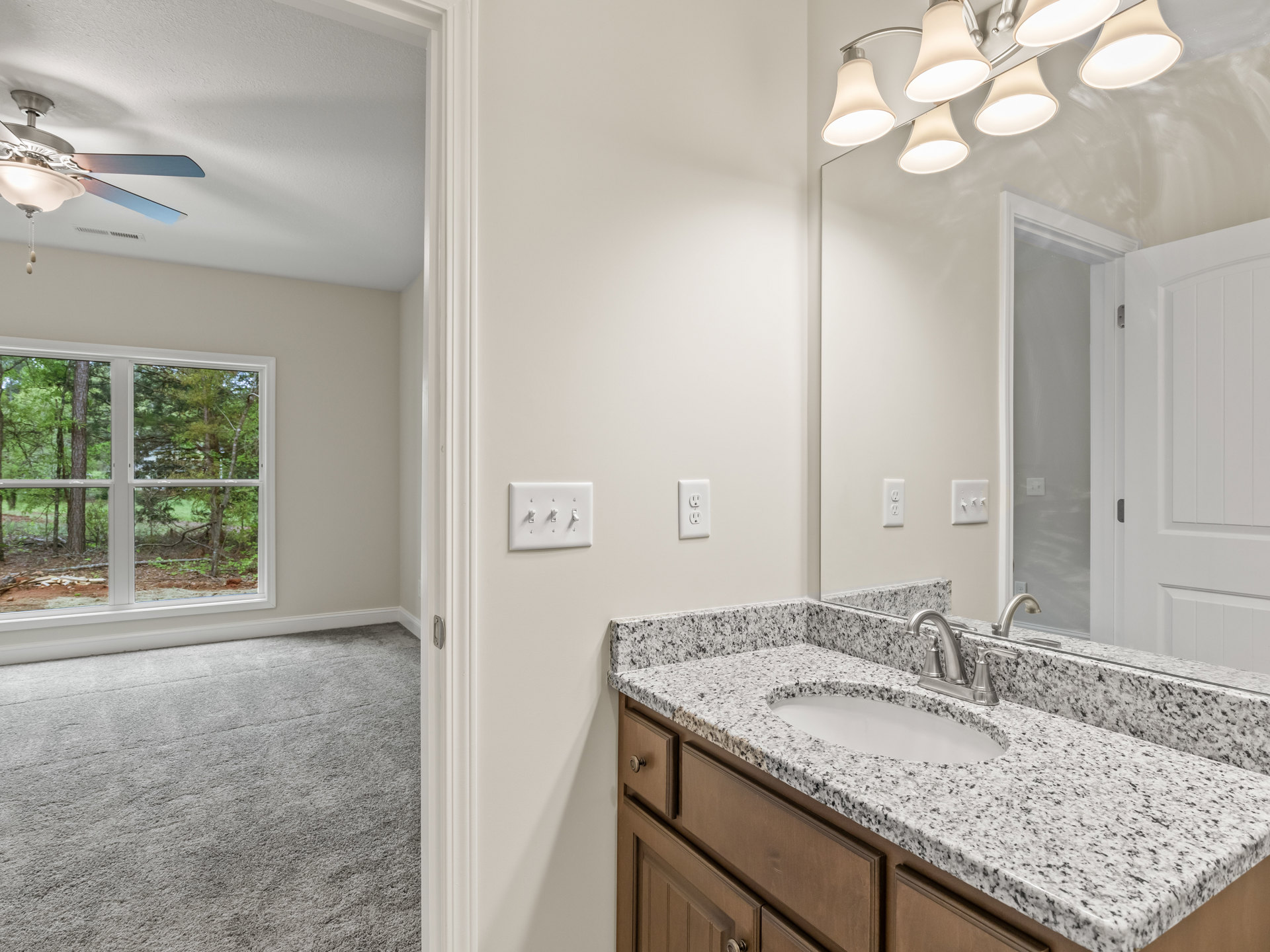Bathroom with marble countertop, undermount sink, silver faucet, large framed mirror with mounted lights, tile backsplash, cabinetry, and window overlooking trees
