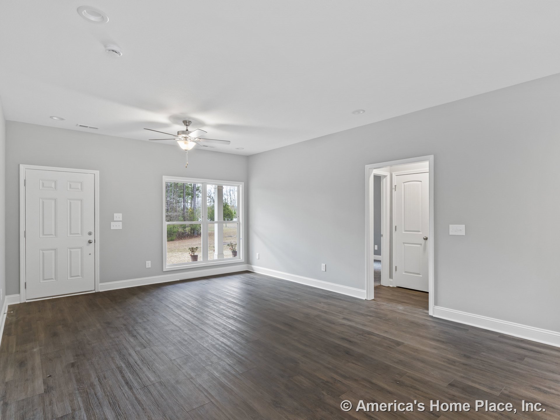 Wide plank wood flooring, large double window with white trim, ceiling fan with light, recessed ceiling lights, gray painted walls, white baseboards, entry and interior doors, open
