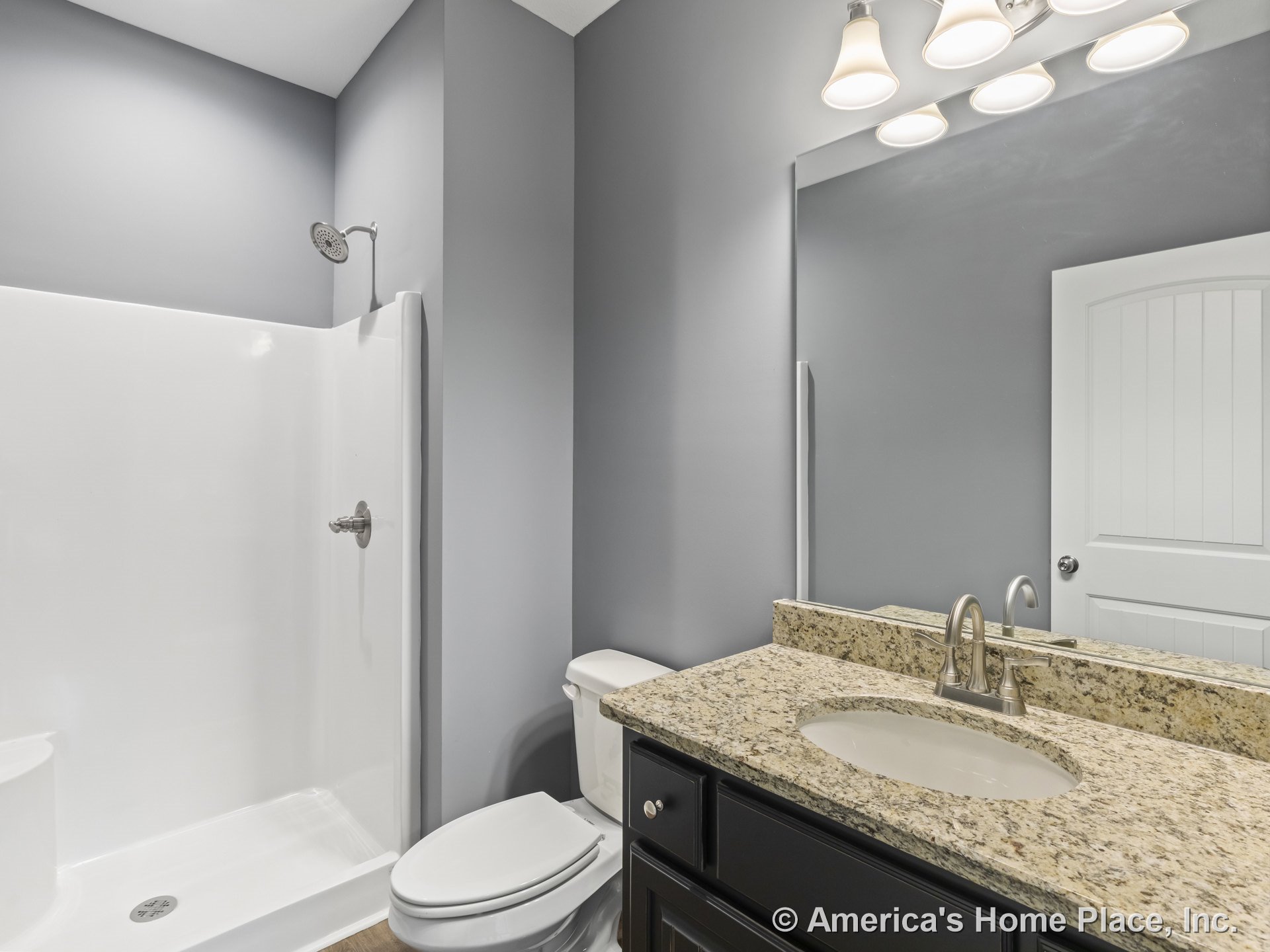 Granite countertop vanity with undermount sink and brushed nickel faucet, large wall mirror topped by three-light fixture, walk-in shower featuring built-in seat and brushed nickel