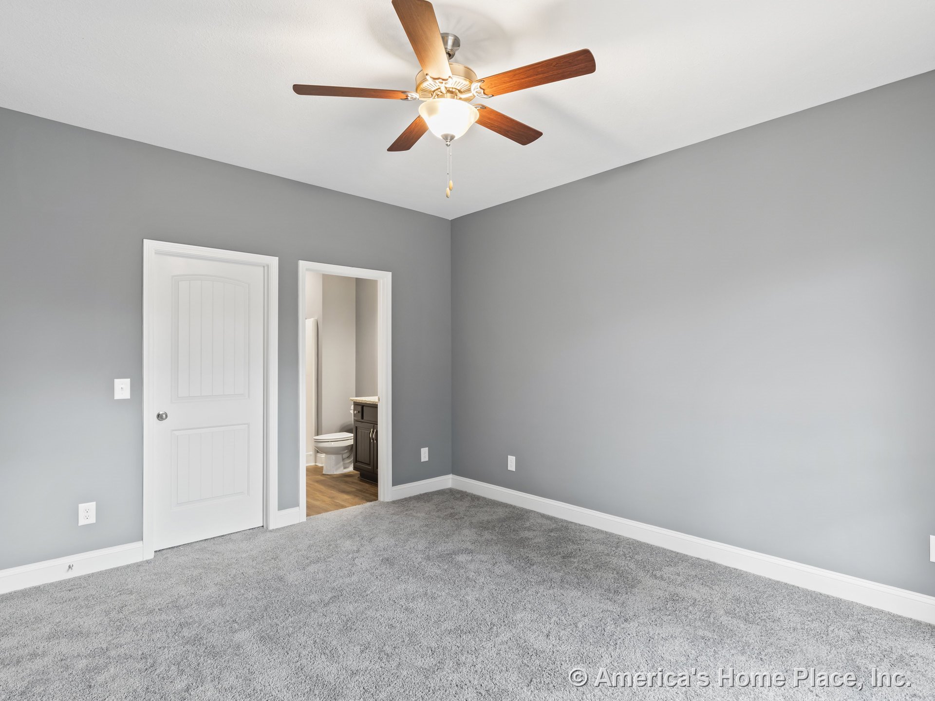 Bedroom with gray carpet flooring, gray walls, white trim and baseboards, paneled interior door, ceiling fan with light fixture, and bathroom entry showing vanity; neutral color
