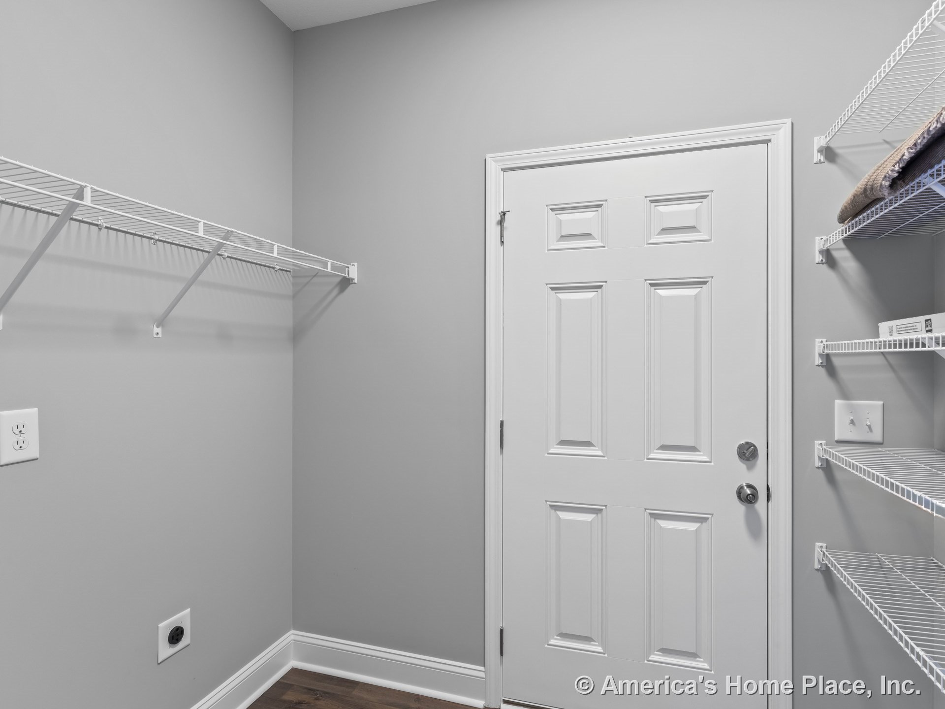 Laundry room with gray painted walls, wire wall shelving, wood-look flooring, white paneled door, baseboard trim, and utility outlet.