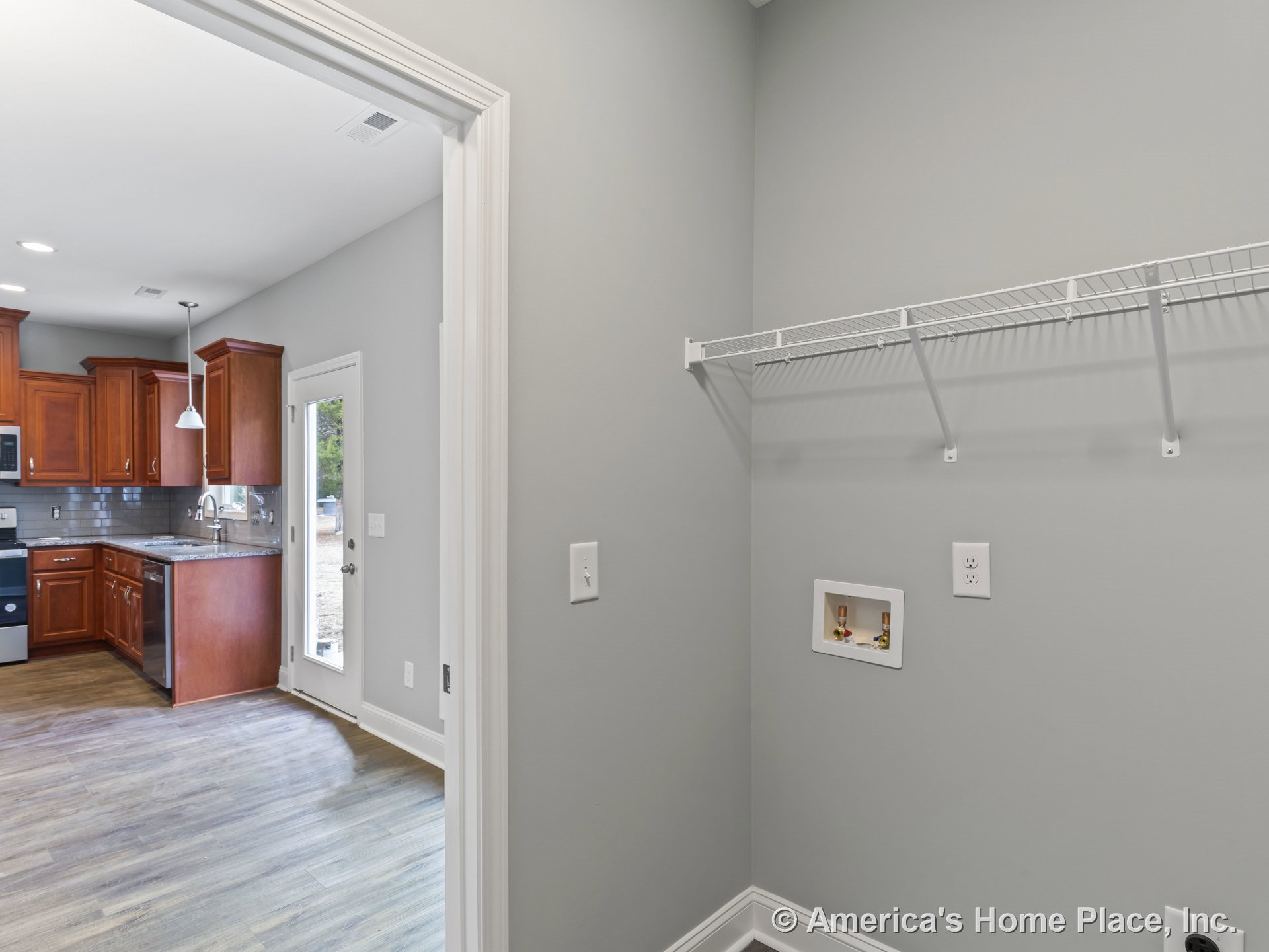 Laundry room with wire shelving and hookups, open doorway leading to kitchen with wood cabinetry, granite countertops, tile backsplash, pendant lighting, glass exterior door
