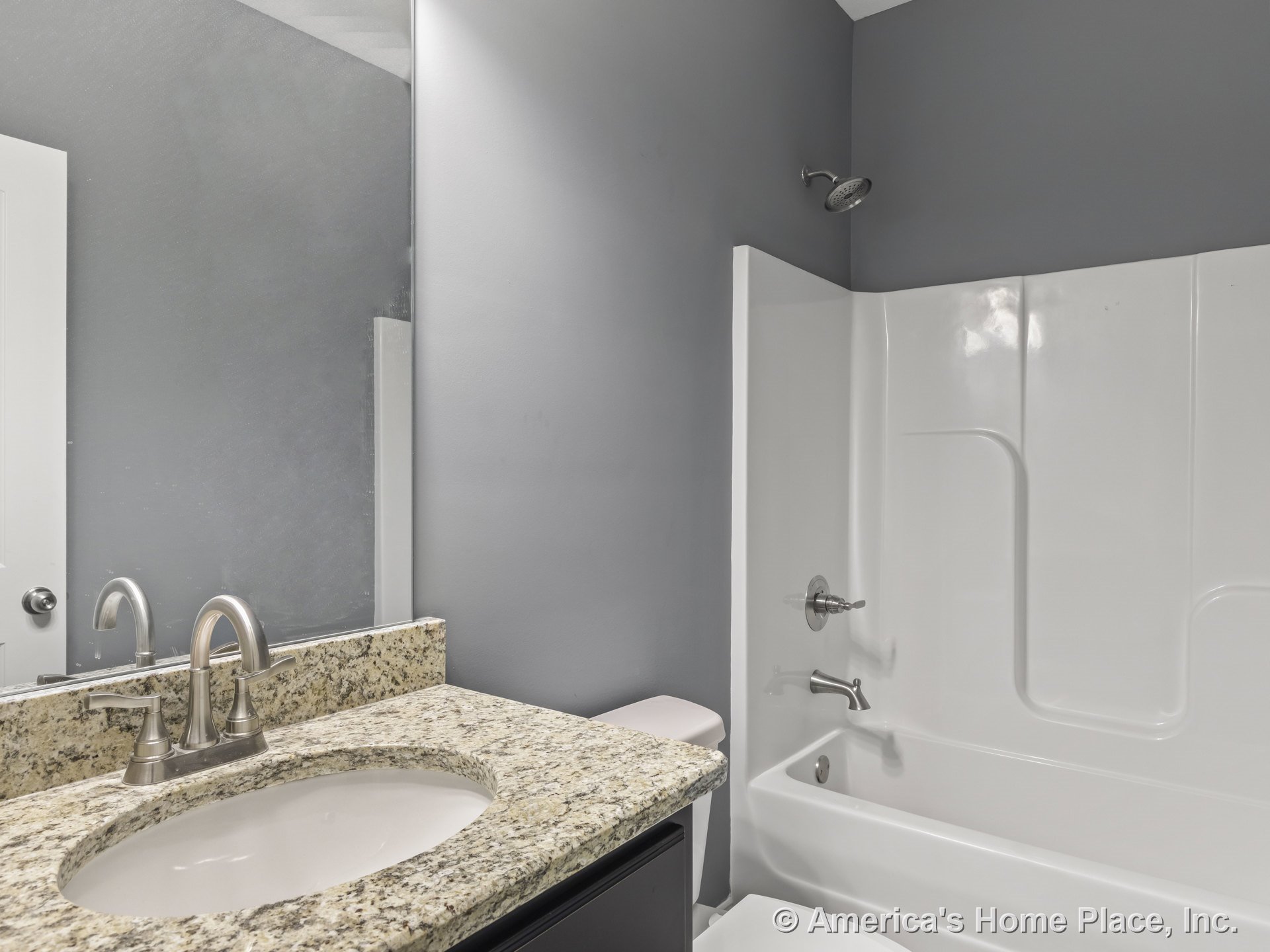 Granite countertop vanity with brushed nickel faucet, large wall mirror above, built-in fiberglass tub and shower, gray painted walls, white trim and door.