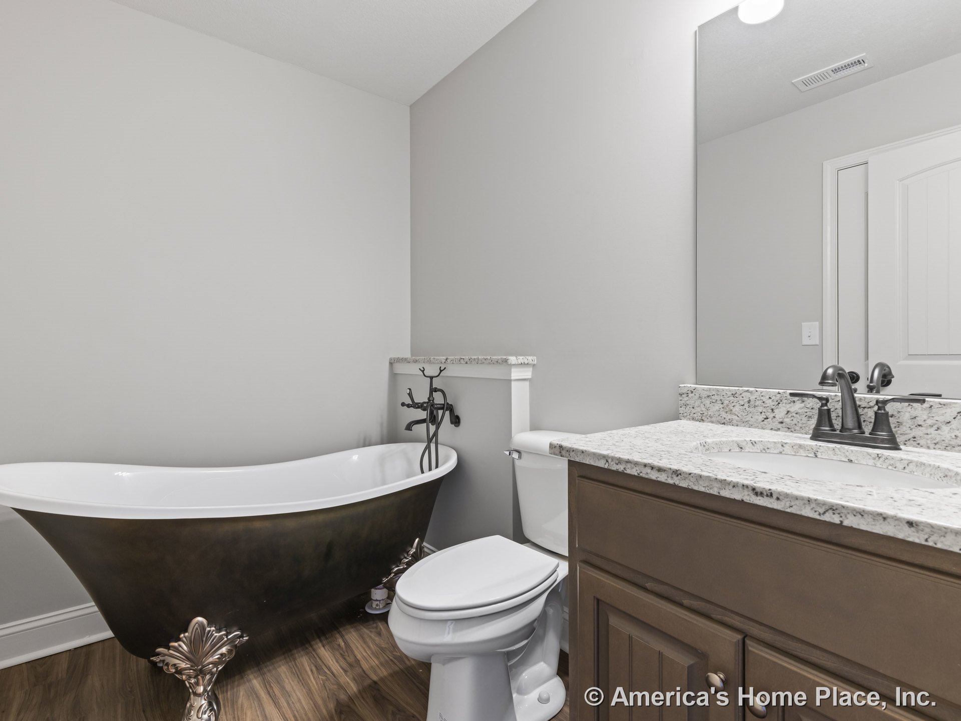 Freestanding clawfoot bathtub beside granite vanity with single undermount sink, wall-mounted faucet, dark wood cabinetry, wood-look flooring, large mirror, neutral walls, and