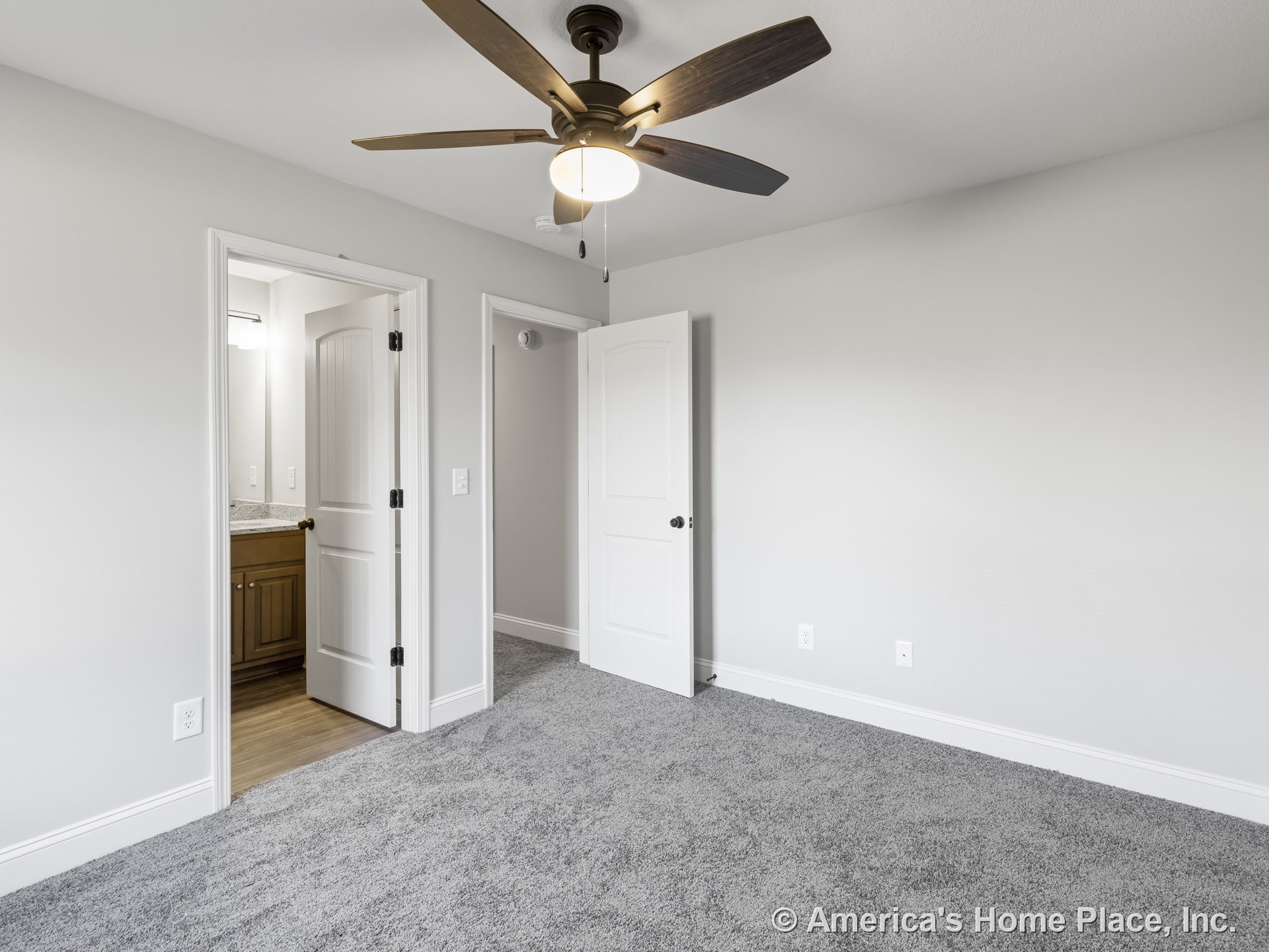 Bedroom with gray carpet flooring, white paneled doors, ceiling fan with wooden blades and light fixture, neutral wall color, white baseboards and trim; adjacent bathroom features