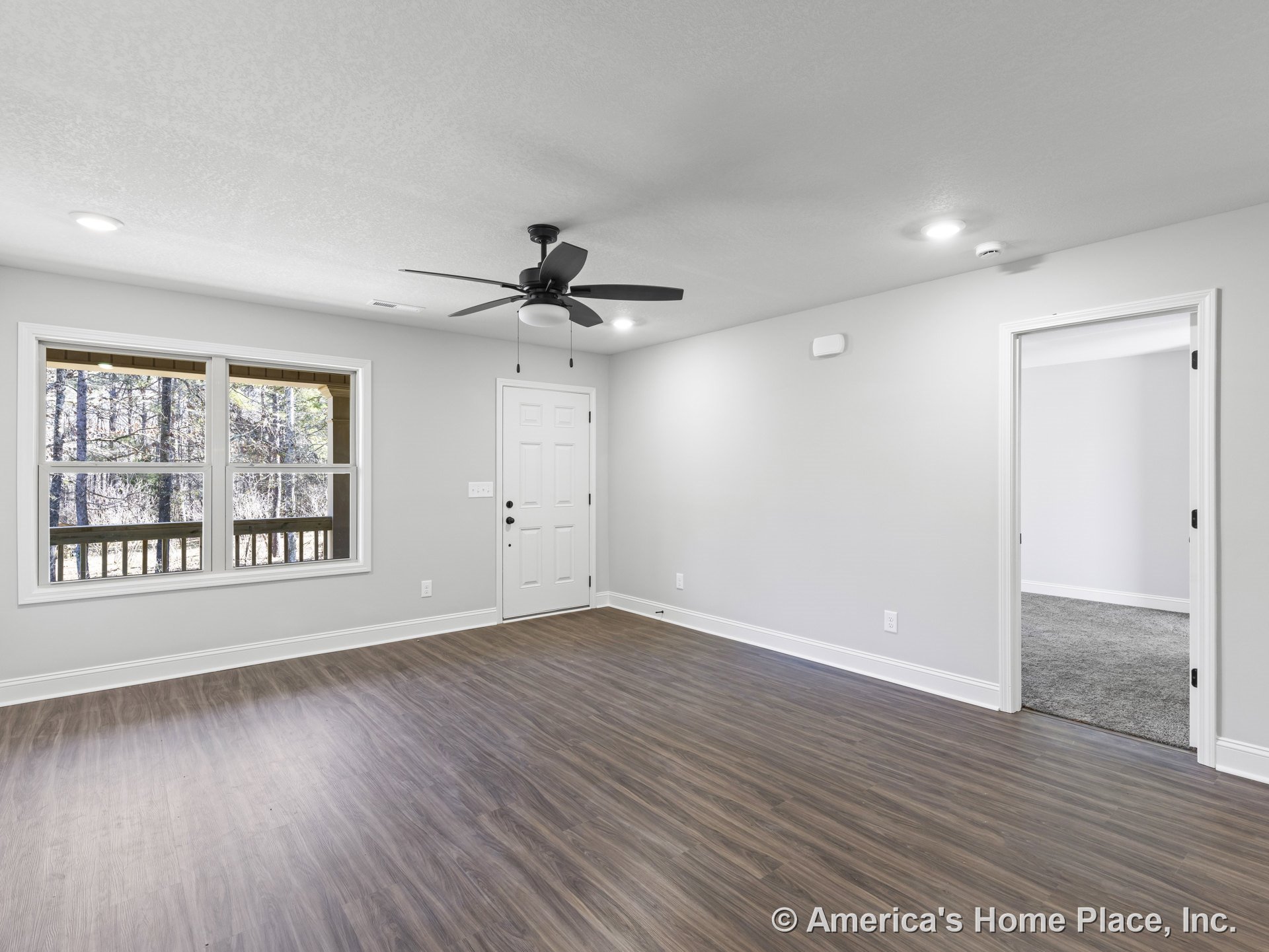 Living room with wood-look vinyl plank flooring, recessed ceiling lights, ceiling fan with light, large window framed in white trim, panel-design front door, neutral walls with