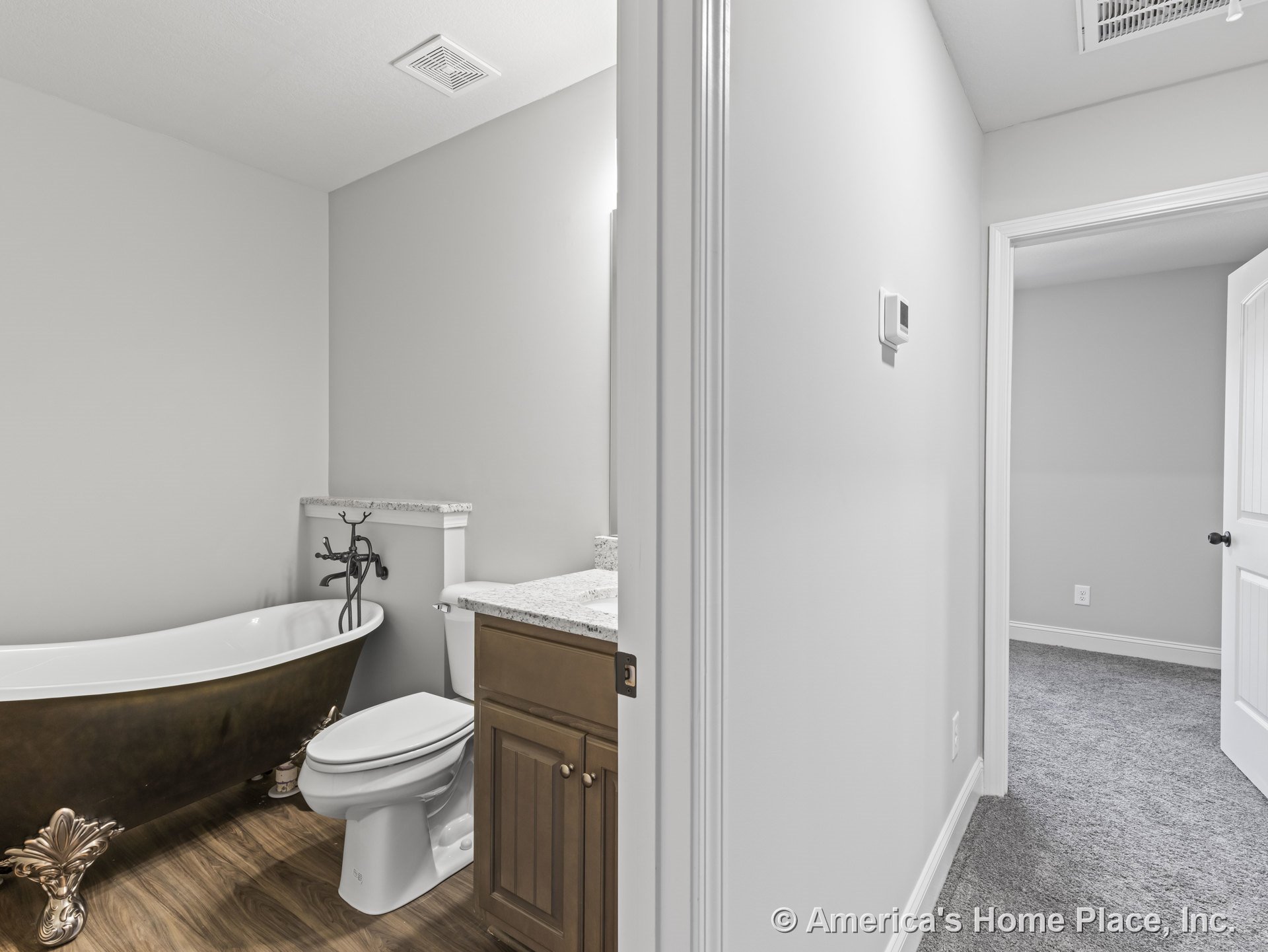 Clawfoot bathtub beside granite vanity countertop and wood cabinetry, white toilet, vinyl plank flooring, and carpeted hallway visible through open doorway.